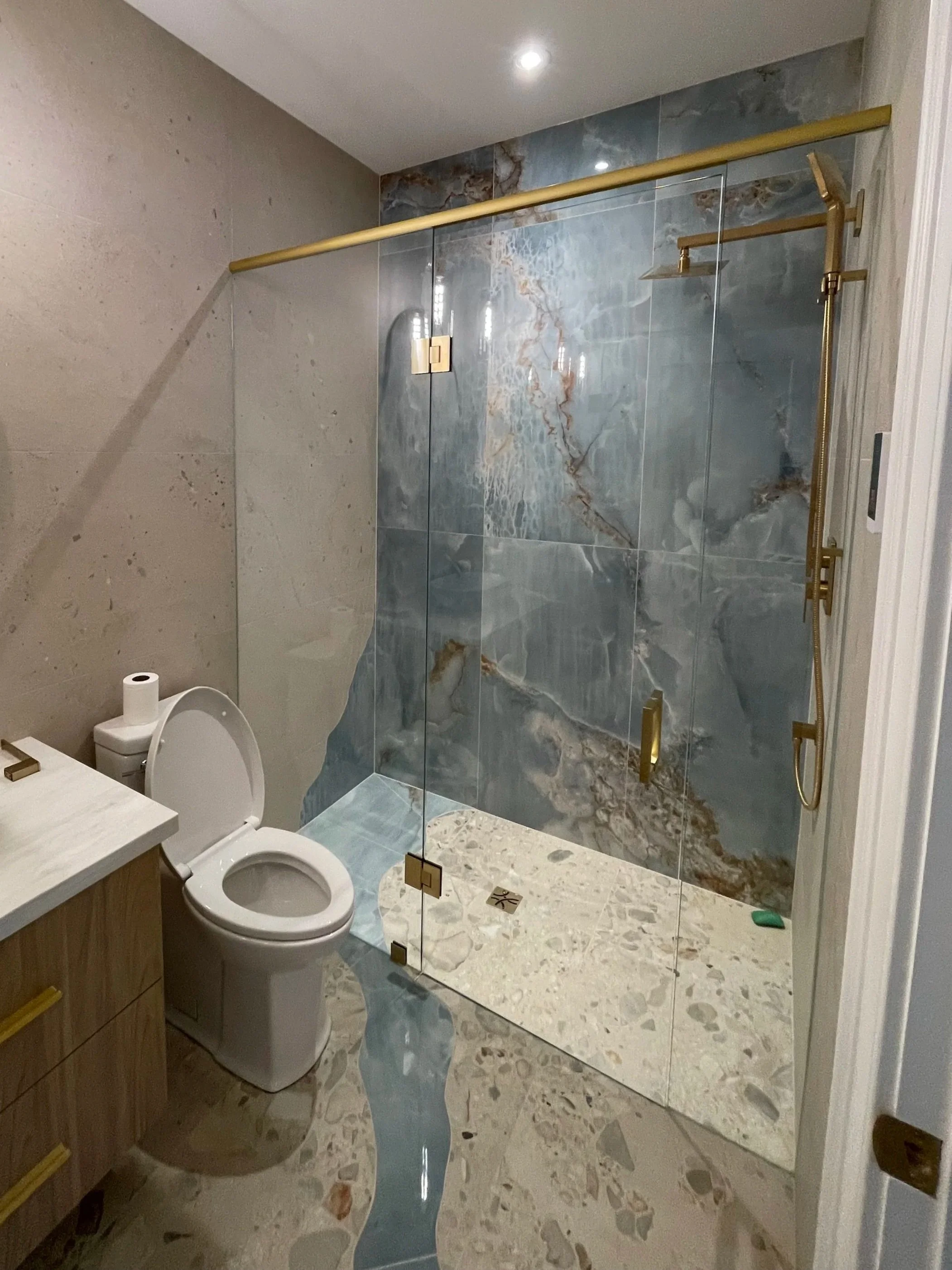 Modern bathroom with a glass shower enclosure, custom blue marble tile walls mimicking the movement of water, gold fixtures, a white toilet, and terrazzo floors.  Tile Work and Contractor Kawartha Lakes, GTA, Muskokas. Bathroom renovation
