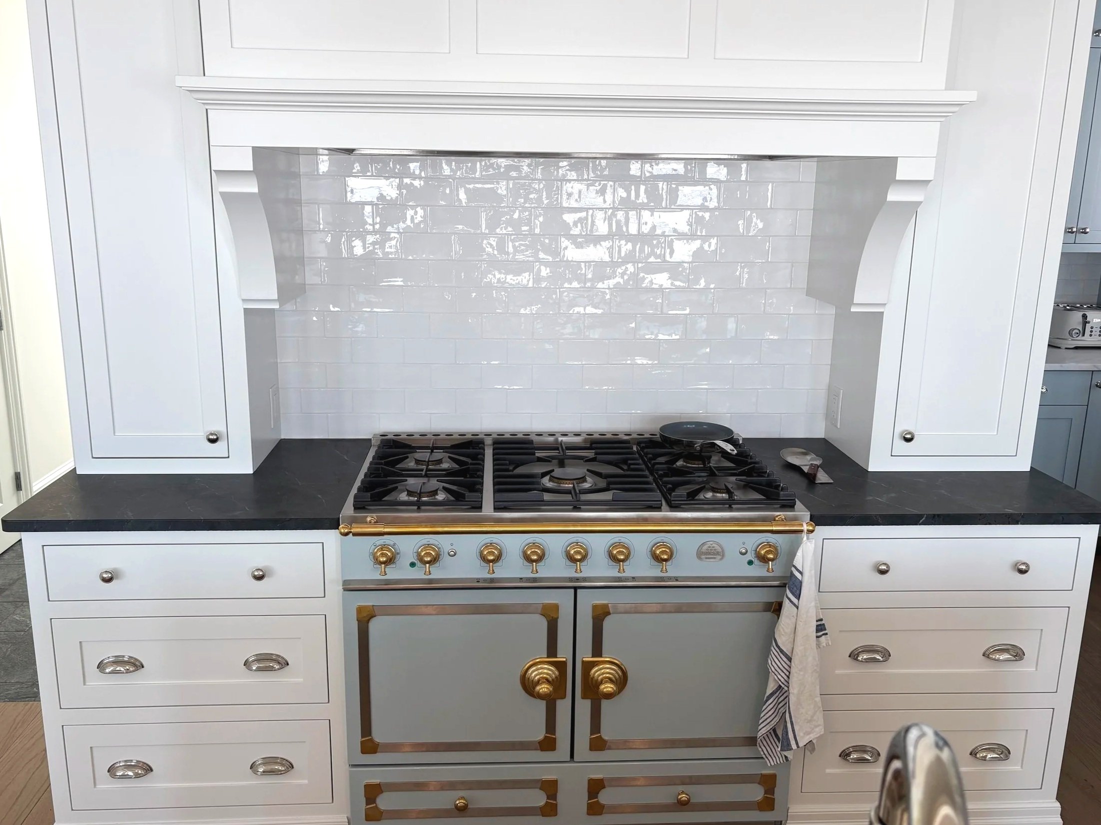 A modern kitchen with a white color scheme, black countertop, and a stainless steel gas range with gold accents. There are white cabinets above and below the counter, and a white subway tile backsplash.  Tile Work and Contractor Kawartha Lakes, GTA,