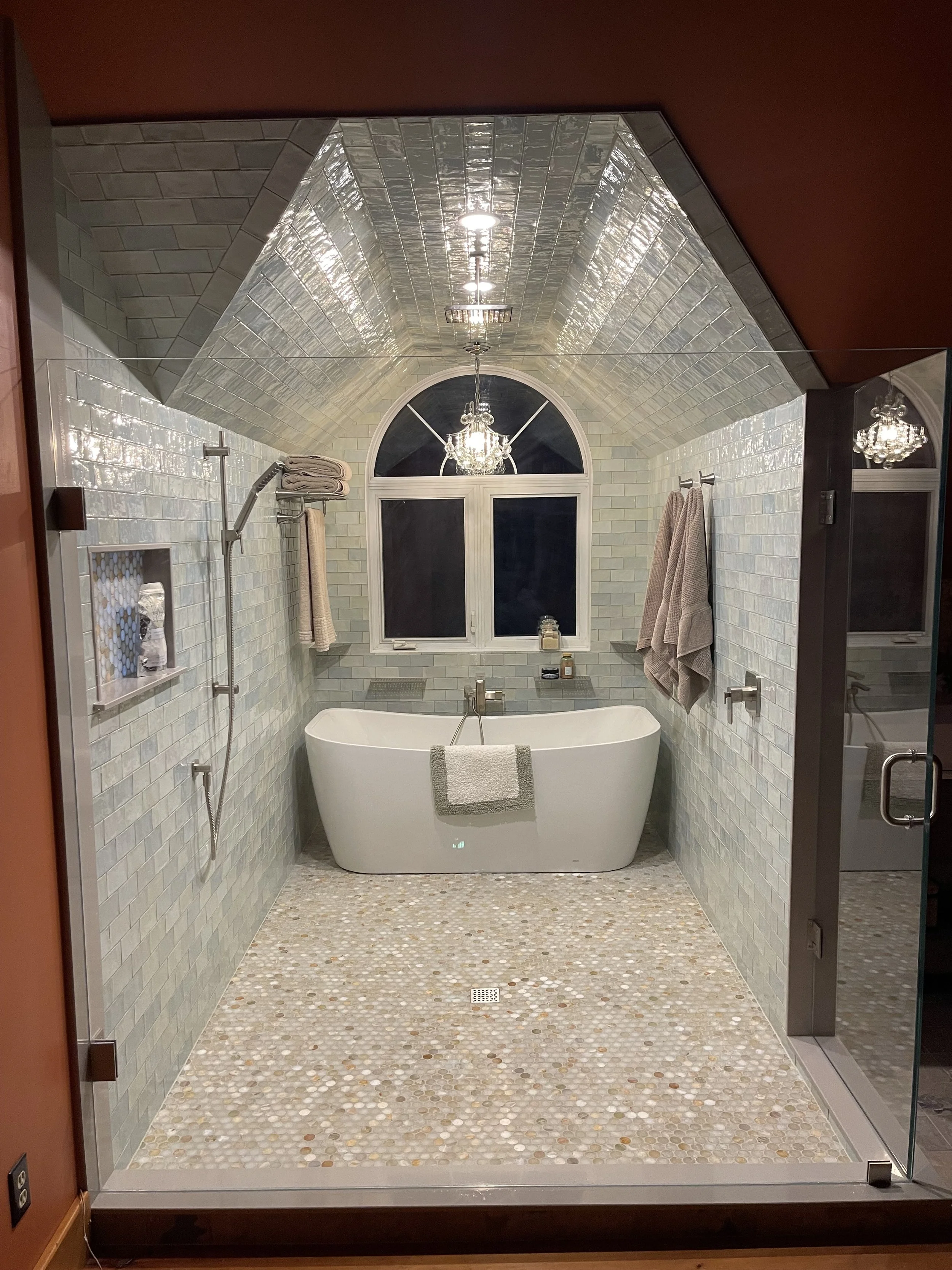 Modern bathroom with a freestanding bathtub in front of a large arched window, tiled walls, pendant chandelier lighting, and towels hanging on hooks.  Tile Work and Contractor Kawartha Lakes, GTA, Muskokas.