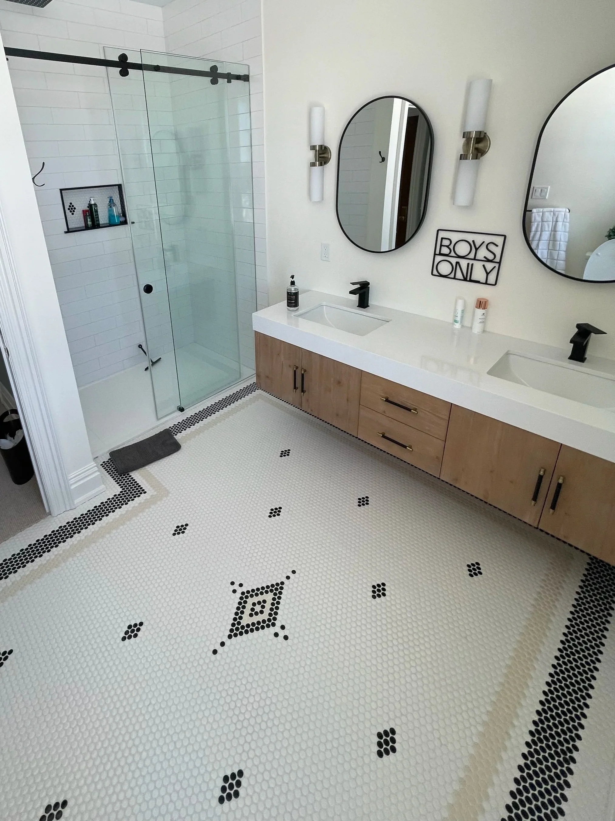 Modern bathroom with dual sinks, wooden vanity, two mirrors, wall-mounted lights, a glass shower enclosure with a detailed, patterned tile floor. Tile Work and Contractor Kawartha Lakes, GTA, Muskokas.