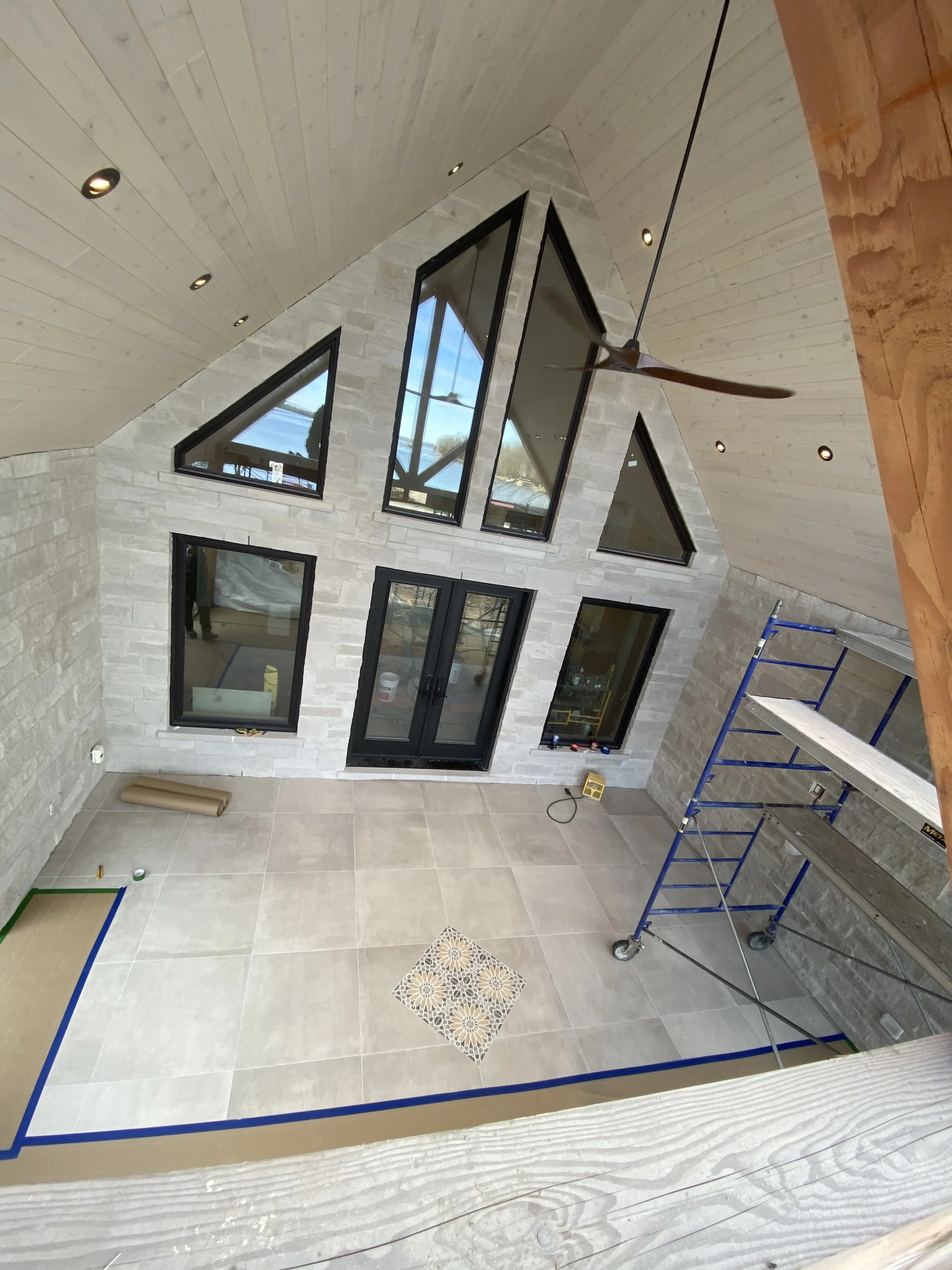 Interior view of a room under construction with stone walls, large black-framed windows, beige tiled flooring, a blue scaffold, a yellow electric heater, a ceiling fan, and a vaulted wooden ceiling with recessed lighting.  Tile Work and Contractor 