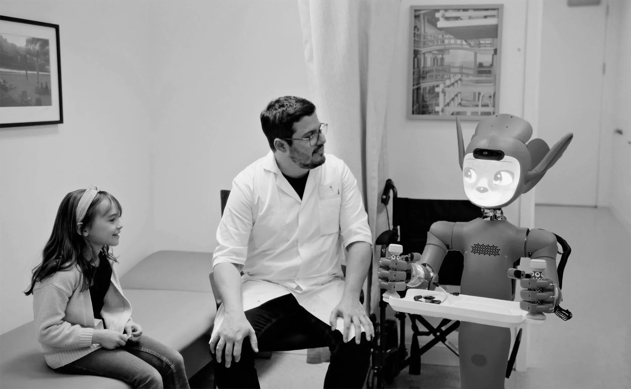 A man and a young girl sit in a room with white walls, watching a robot with a humanoid face and large ears as it interacts with them.