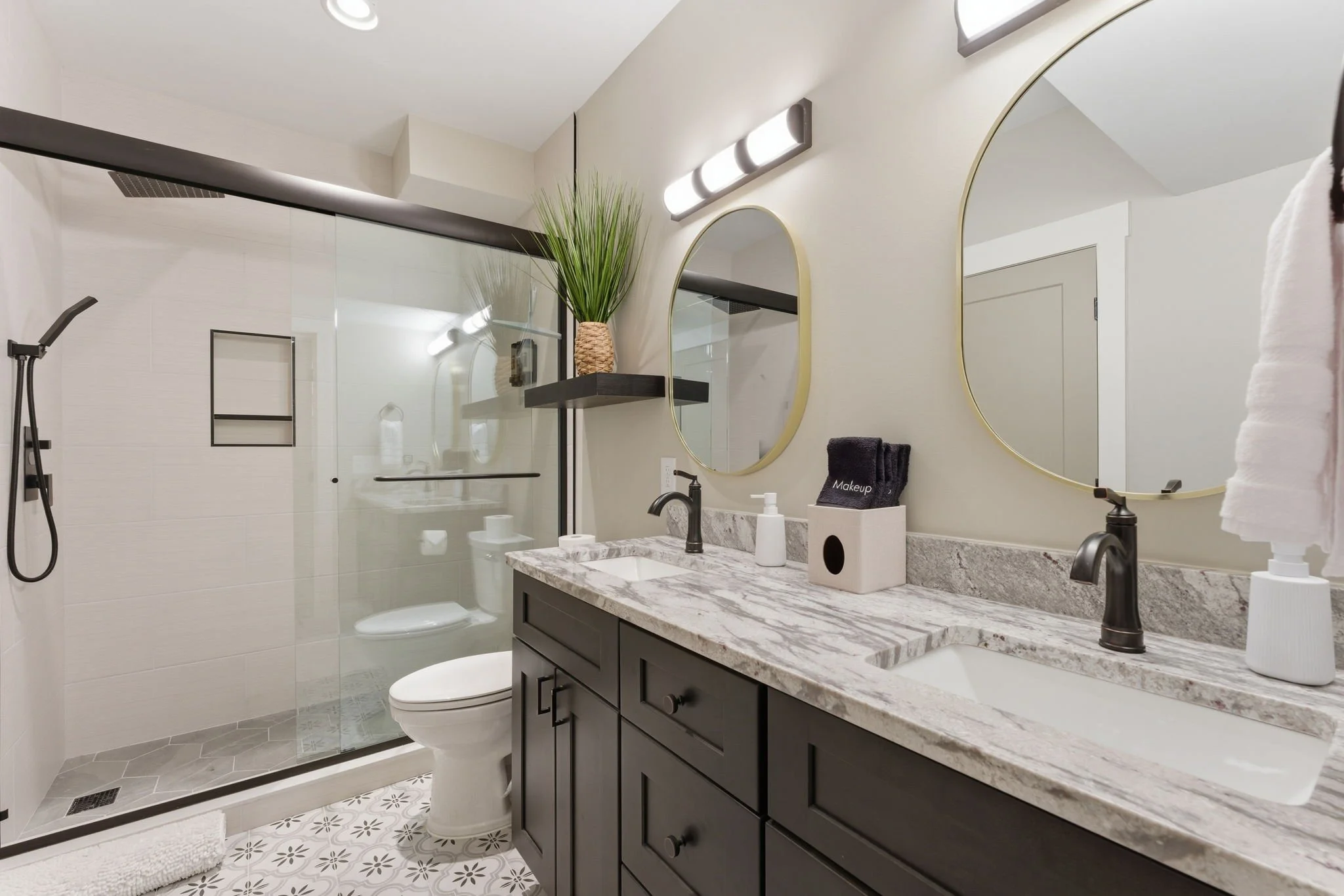 Modern bathroom featuring a double vanity with granite countertop, two oval mirrors, black fixtures, and a glass-enclosed shower with built-in shelves and gray hexagonal tile flooring.