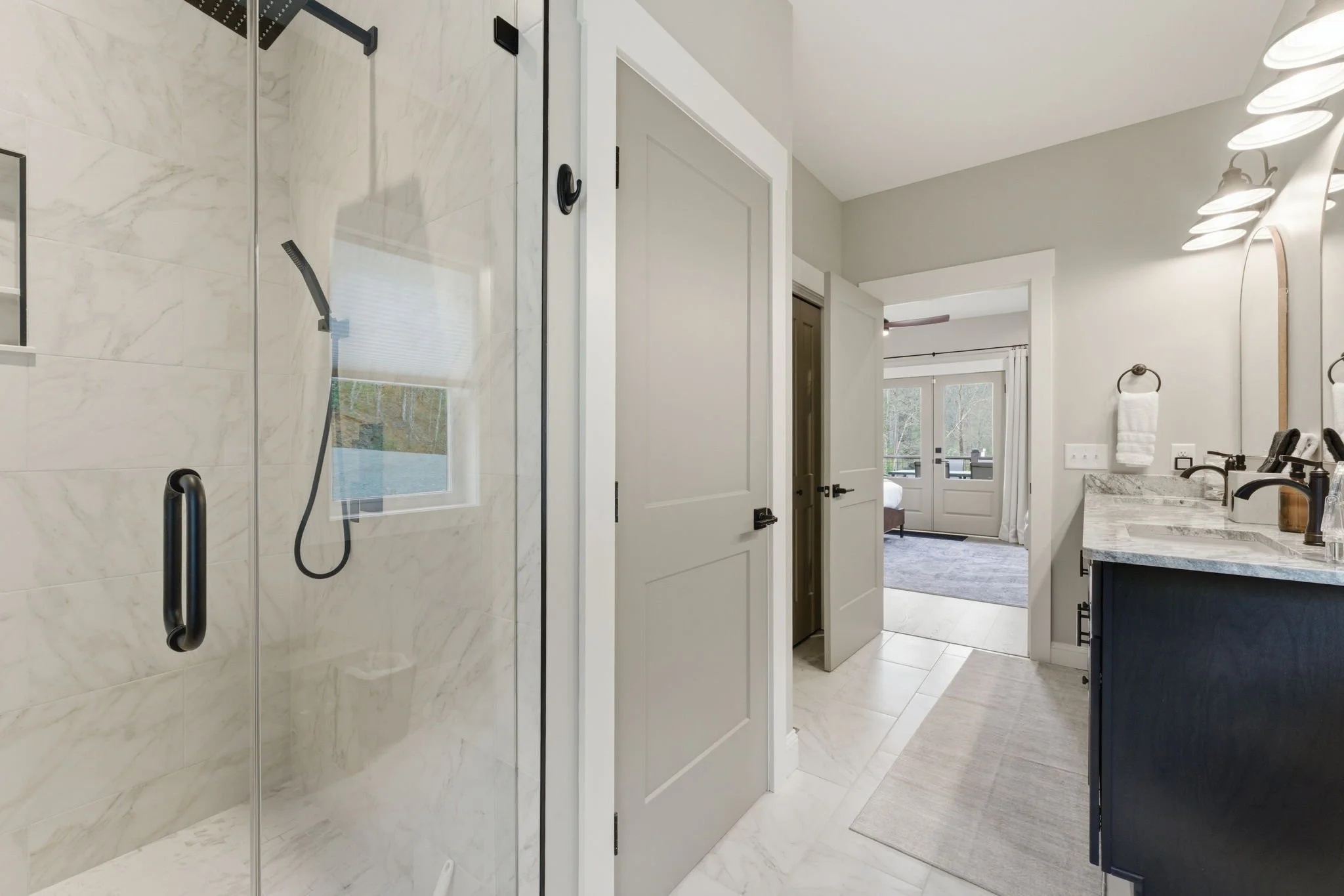 Bathroom with a full tile shower, double vanity, granite countertops, black fixtures, and with a view into a bedroom and living area.