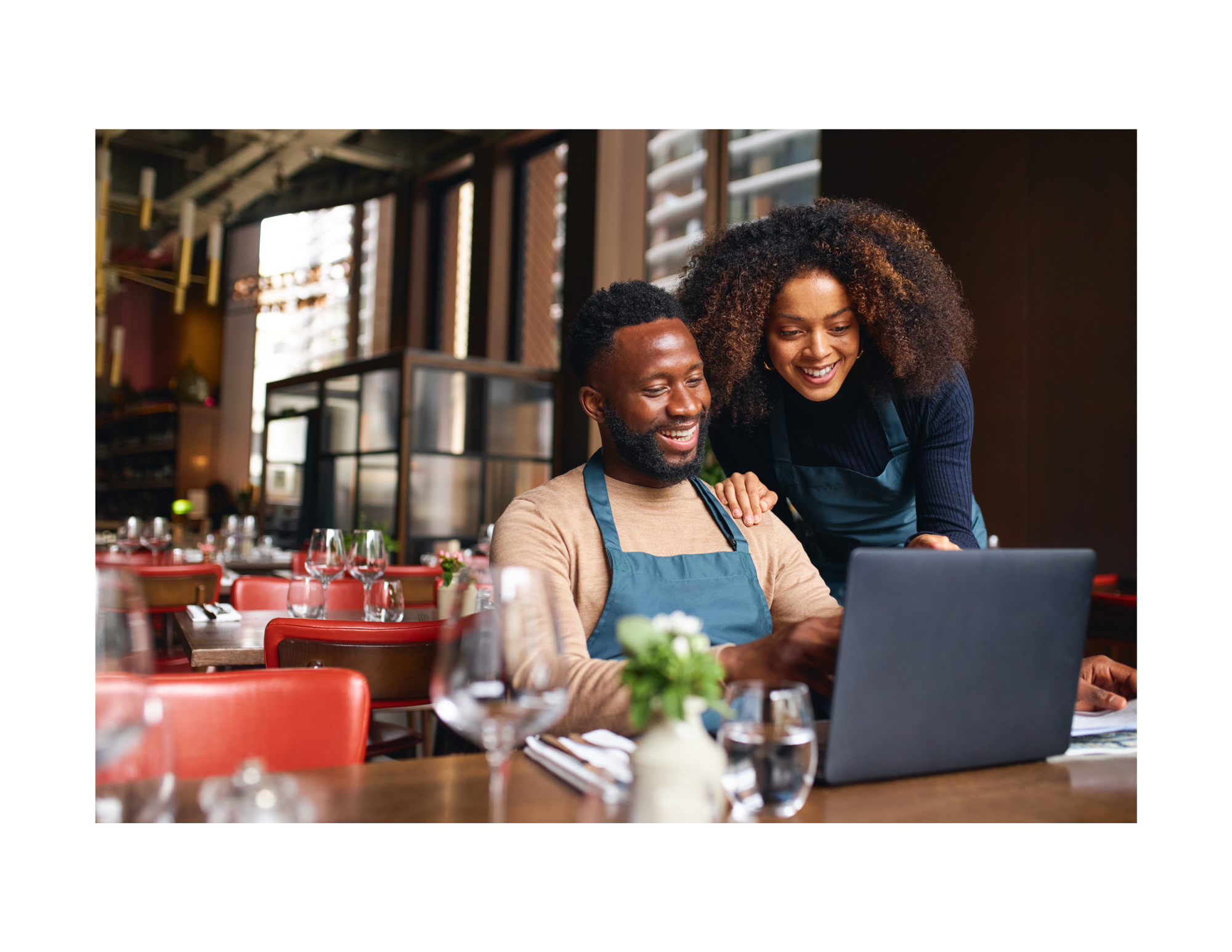 A man and a woman smiling while looking at a laptop in a restaurant. The woman is standing next to the man who is sitting at a table.