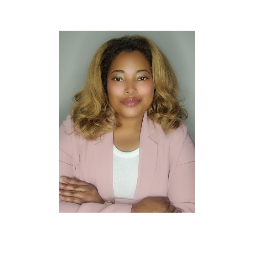 A woman with blonde, wavy hair, wearing a pink blazer and white top, smiling with arms crossed against a gray background.