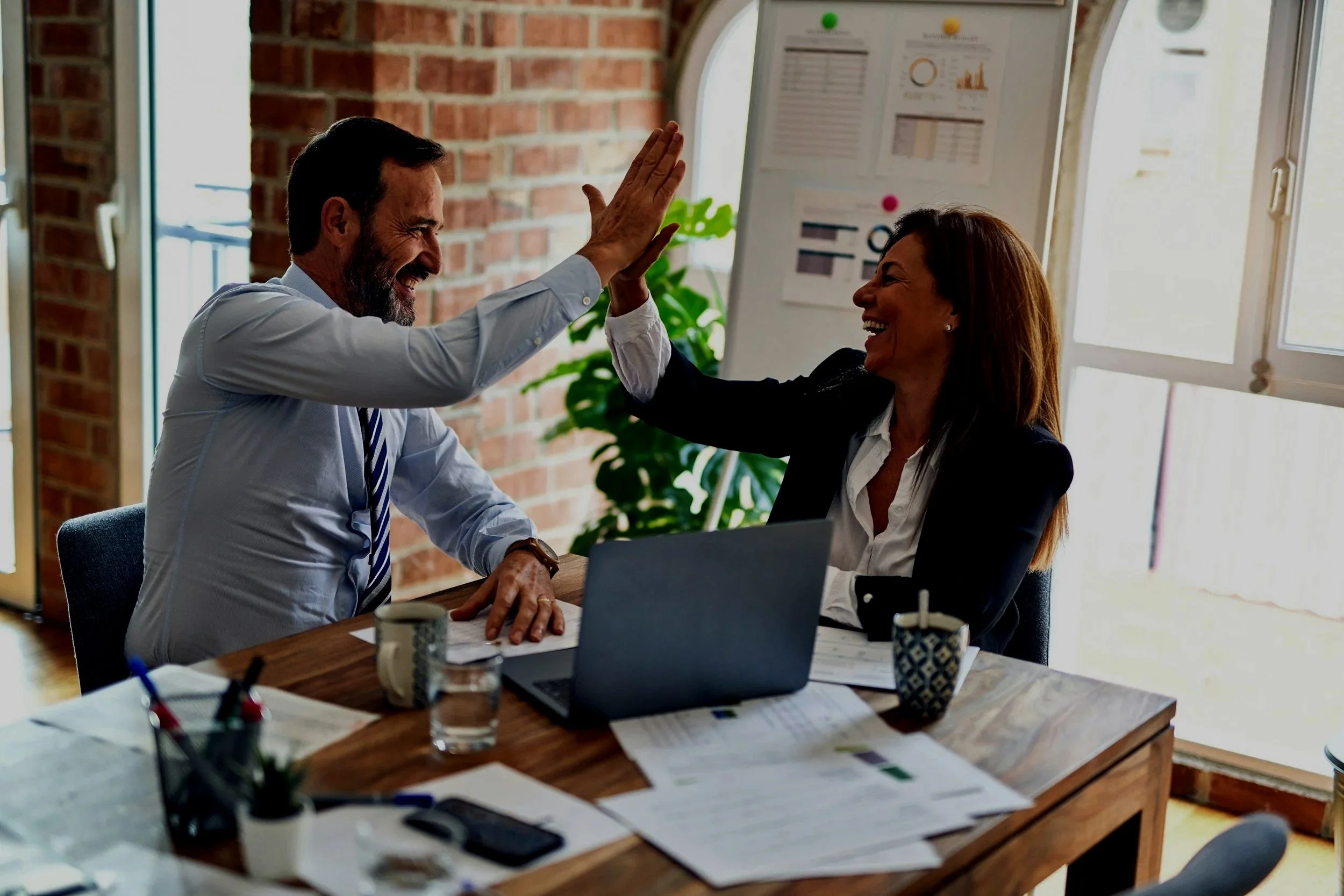 A man and woman in business attire are inside an office, giving each other a high five and smiling. The office has a brick wall, a large window, a desk with papers, a laptop, and cups.