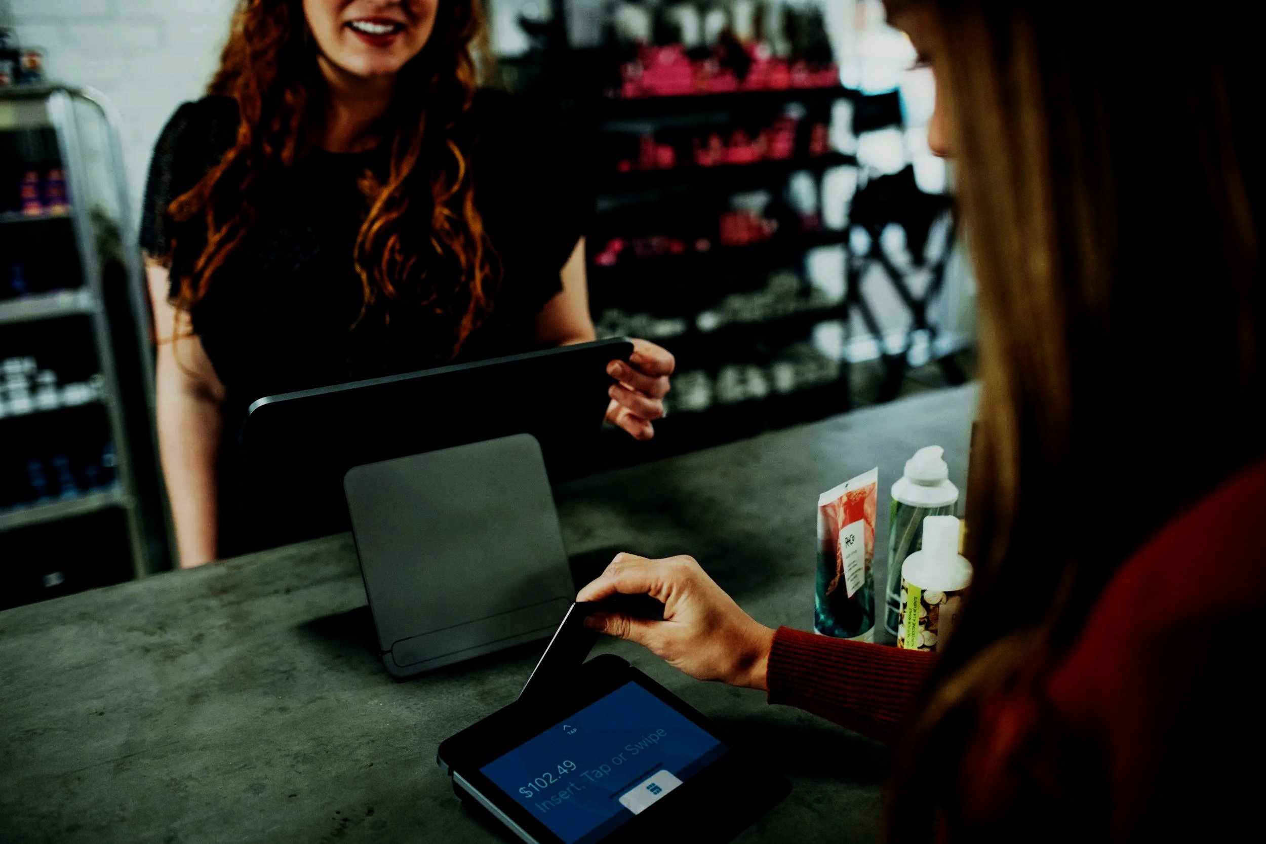 Customer making a contactless payment with a card at a store checkout counter, with a cashier smiling in the background.