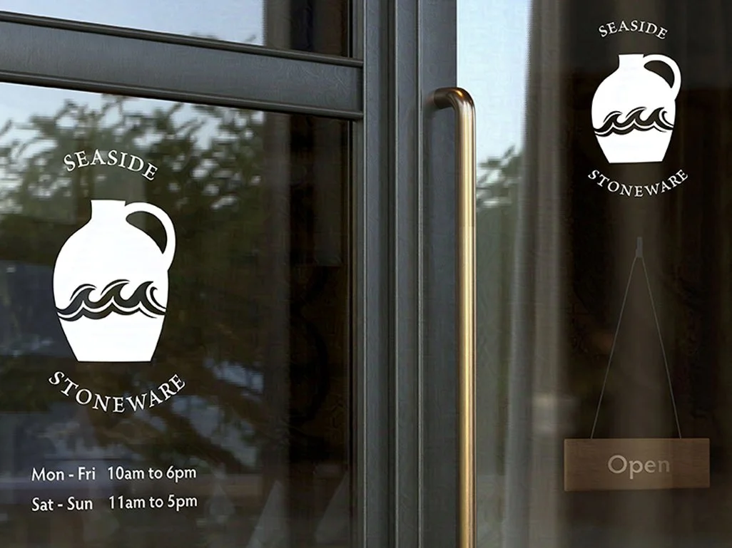 Glass door with a gold handle featuring "SEASIDE STONEWARE" logo of a jug with waves. Hours listed below; an "Open" sign hangs on the right.