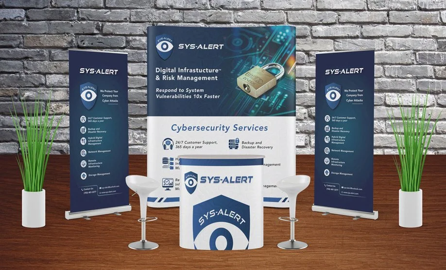 A trade show booth setup for SYS-ALERT features banners and a table with company branding. The backdrop displays a digital padlock graphic and cybersecurity services information. Two modern white chairs are positioned at the table.