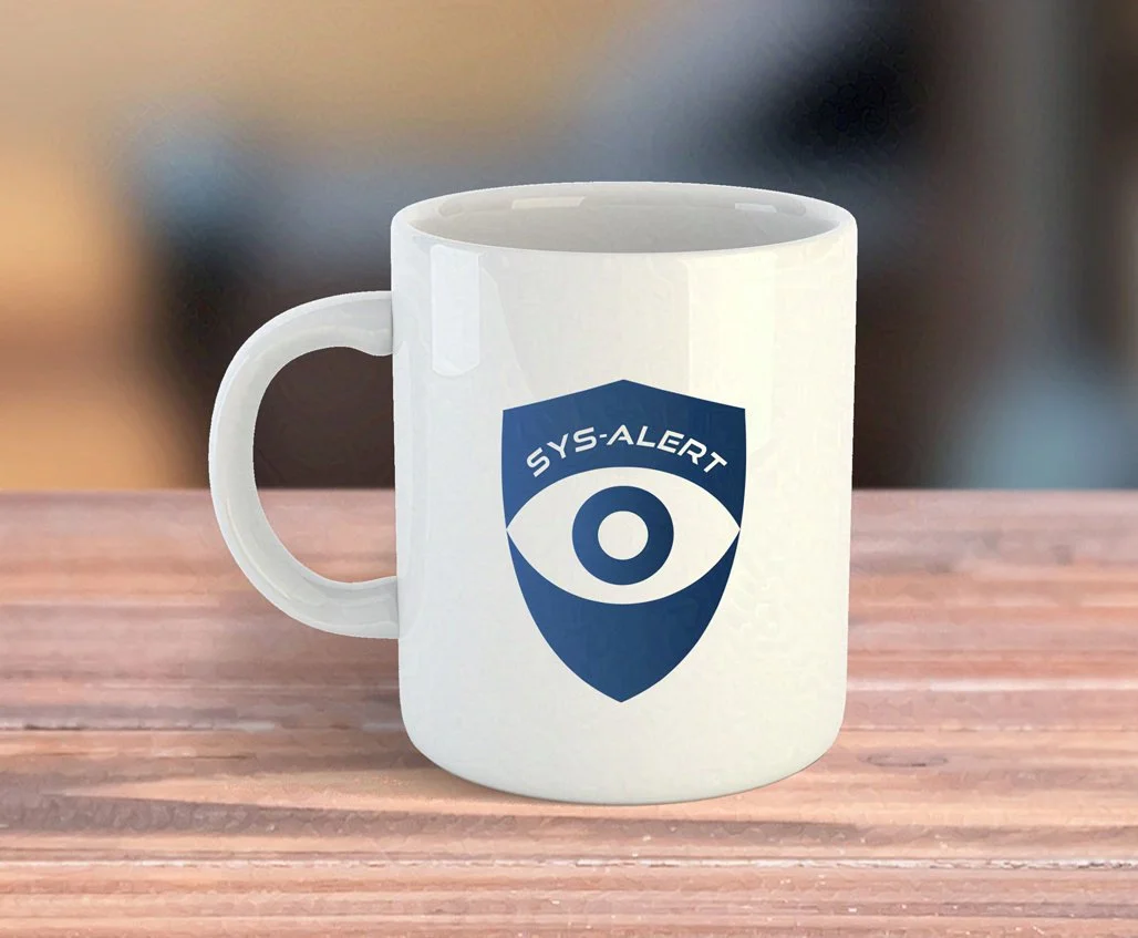 A white coffee mug on a wooden surface features a blue shield logo with an eye graphic and the text SYS-ALERT above the eye. The background is softly blurred.