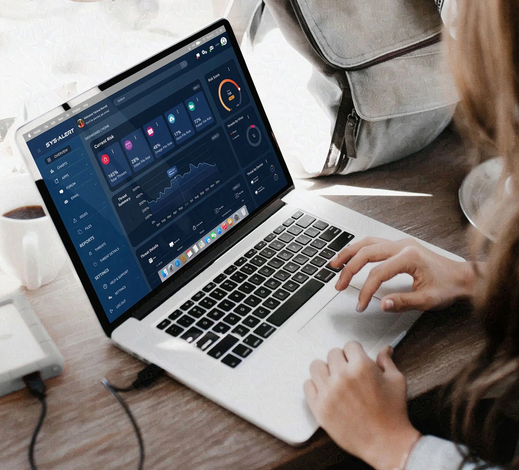 A person using a laptop with a dark-themed dashboard interface displaying charts, graphs, and analytics. A gray bag sits on the wooden table beside the laptop.