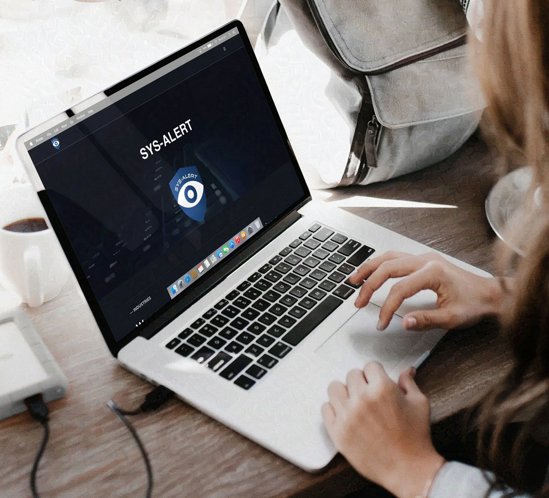 A person types on a laptop displaying a "SYS-ALERT" screen with an eye in a shield symbol. The setting is a well-lit workspace with a coffee cup, conveying focus.