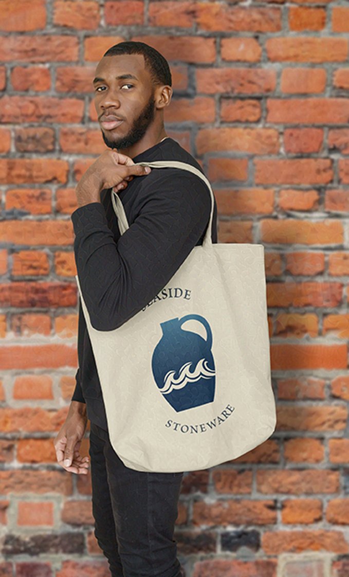 An African American man in a black shirt poses with a beige tote bag featuring a blue stoneware jug and wave design with the words "Seaside Stoneware" against a brick wall.