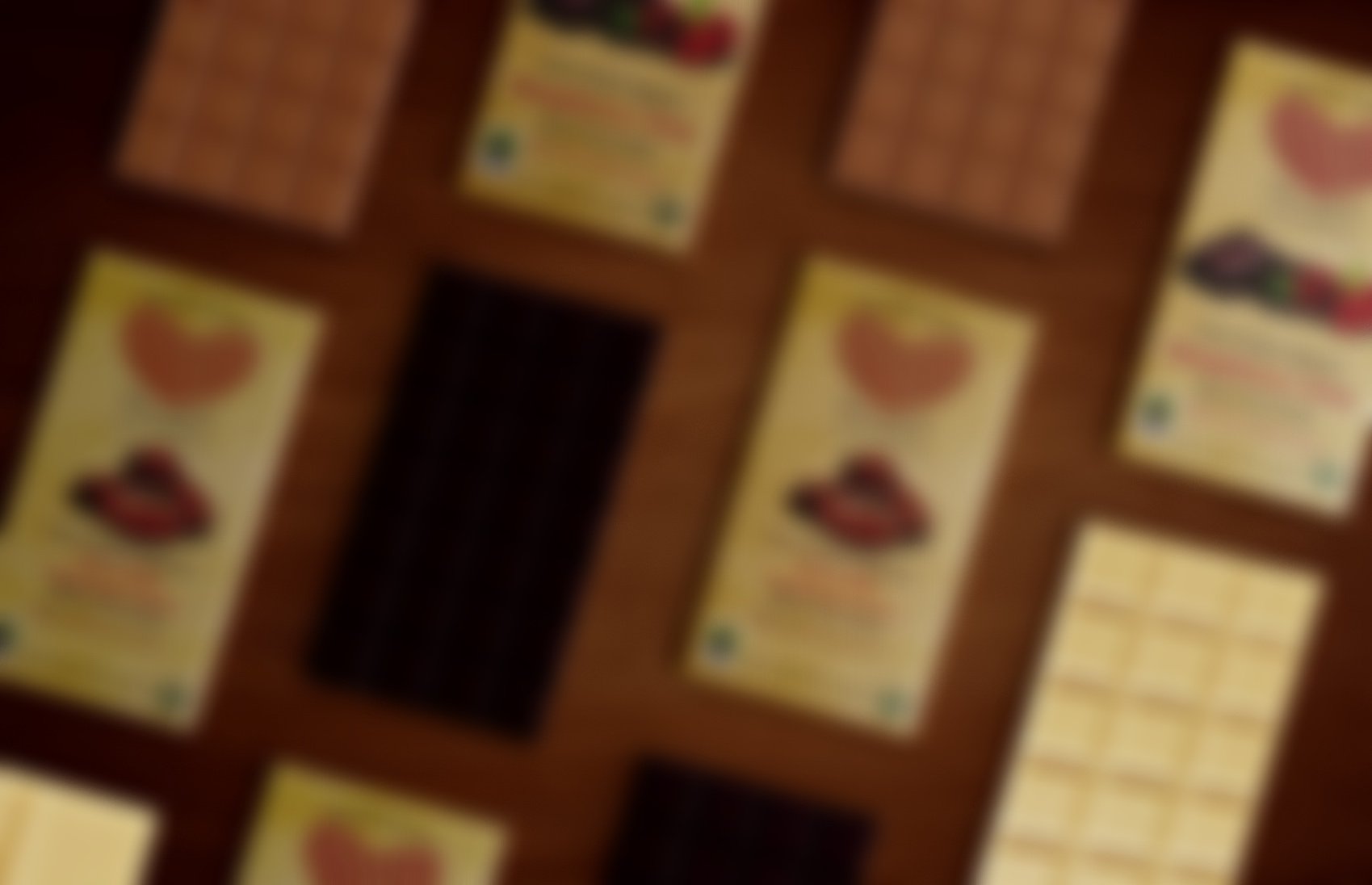 An assortment of chocolate bars and packaging on a wooden surface. Varieties include dark, milk, and white chocolate with vibrant wrappers featuring watercolor illustrations and fair trade labels, conveying a sense of variety and indulgence.