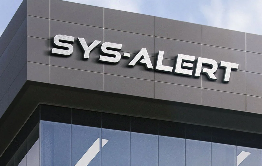 Modern building exterior with large, bold silver letters spelling SYS-ALERT mounted on a dark gray panel above reflective glass windows. The sky is partly visible in the background.