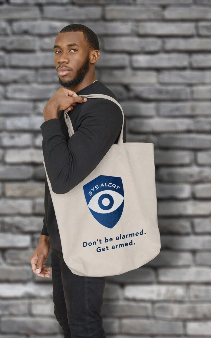 A man in a black outfit stands in front of a gray brick wall, holding a beige tote bag over his shoulder. The bag displays a blue shield logo with an eye and the text, SYS:ALERT Dont be alarmed. Get armed.