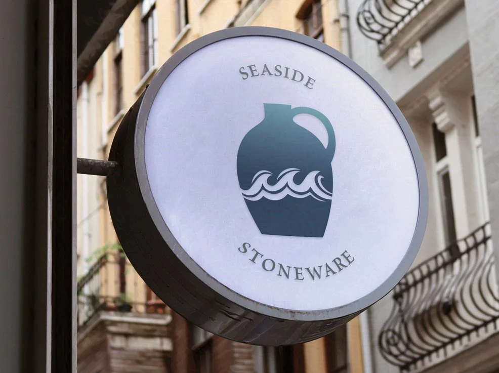 Round sign for Seaside Stoneware featuring a jug silhouette and waves, displayed on a building exterior.