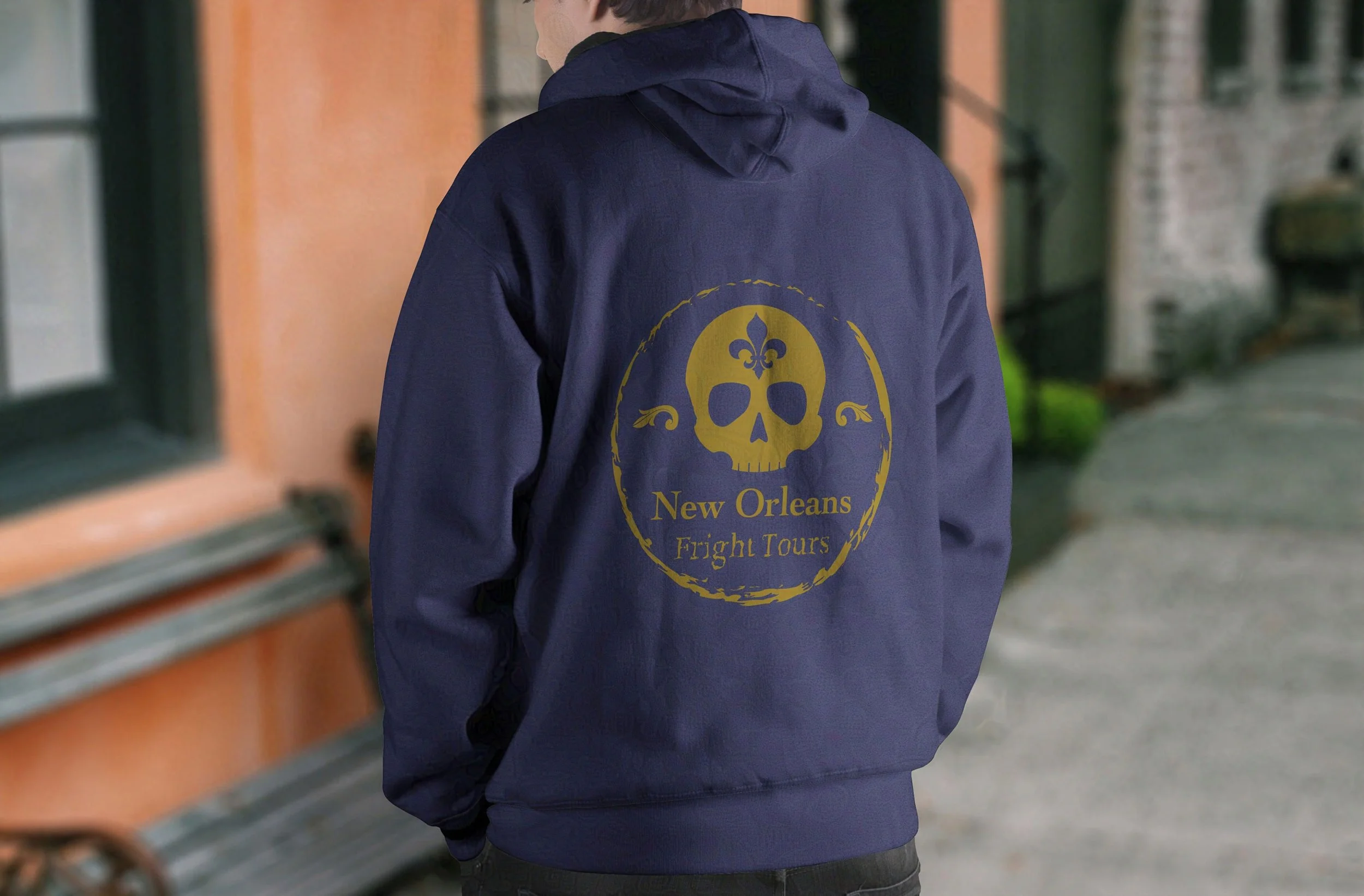 Back view of a white man in a dark purple hoodie with a gold fleur-de-lis “New Orleans Fright Tours” logo, standing on a city street.