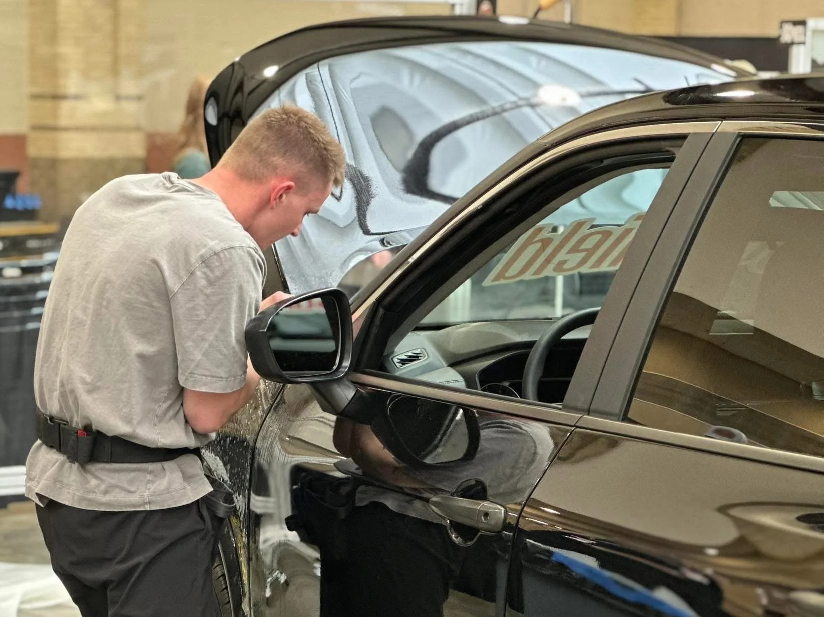 A man is inspecting a black car at a dealership or garage, looking inside the vehicle through the driver's side window.