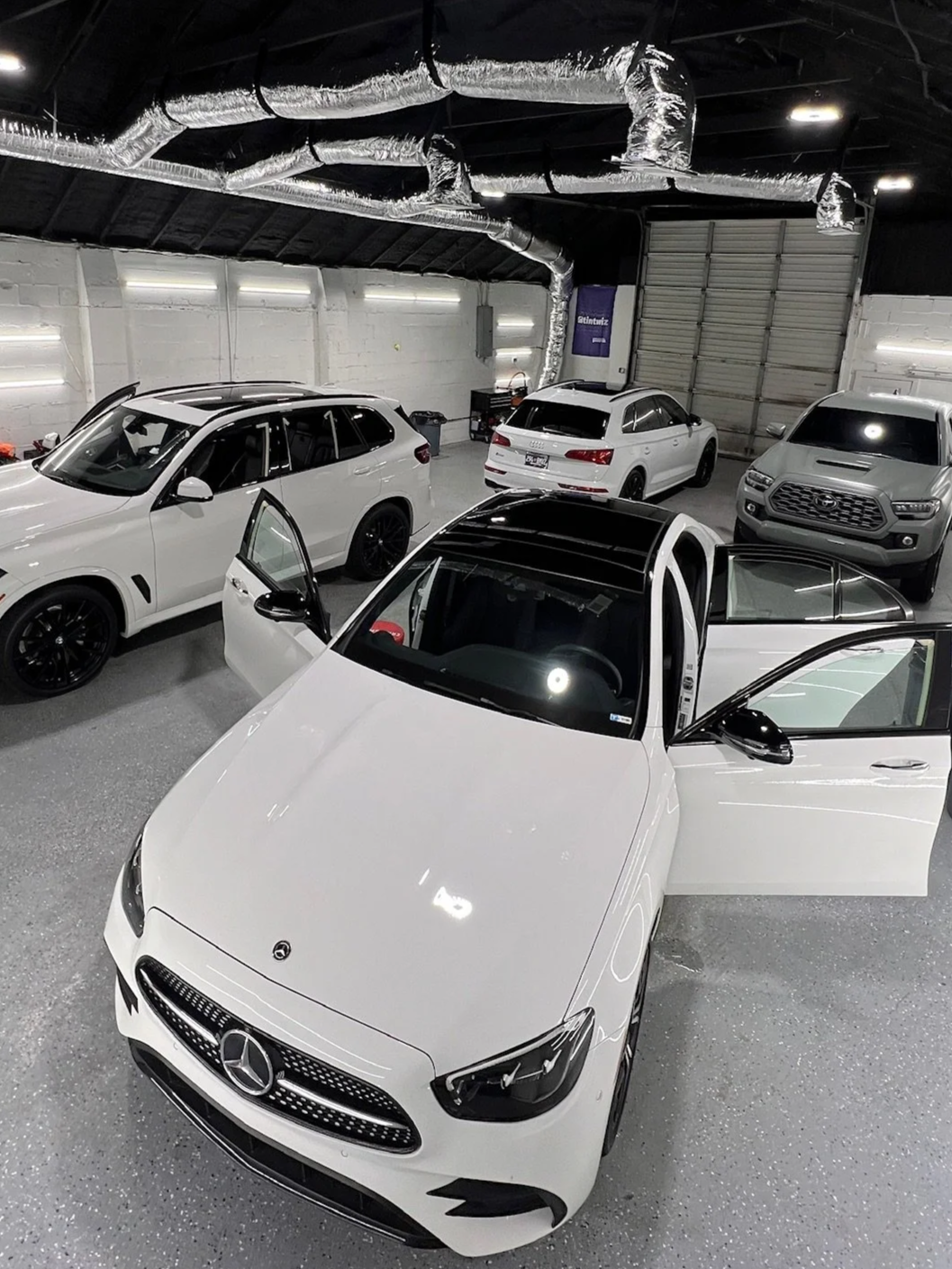 Several white and gray cars in a well-lit indoor garage with exposed ductwork and a large garage door.
