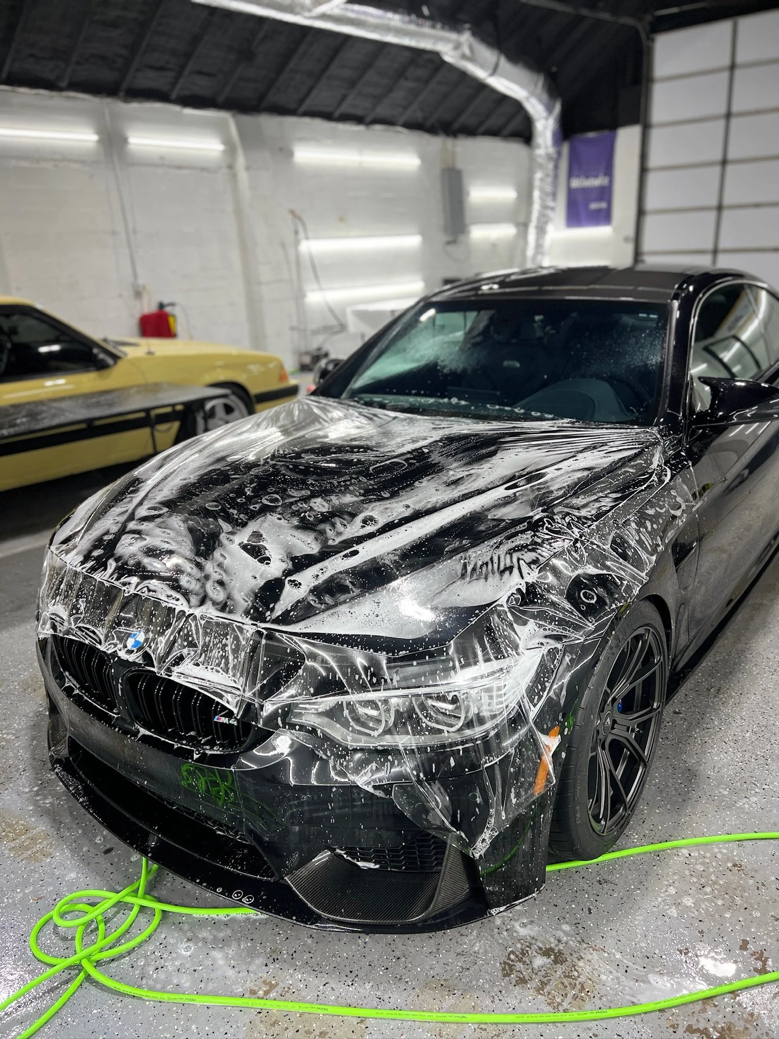 A black BMW M4 car being washed with soap foam in a garage, with a yellow car in the background.