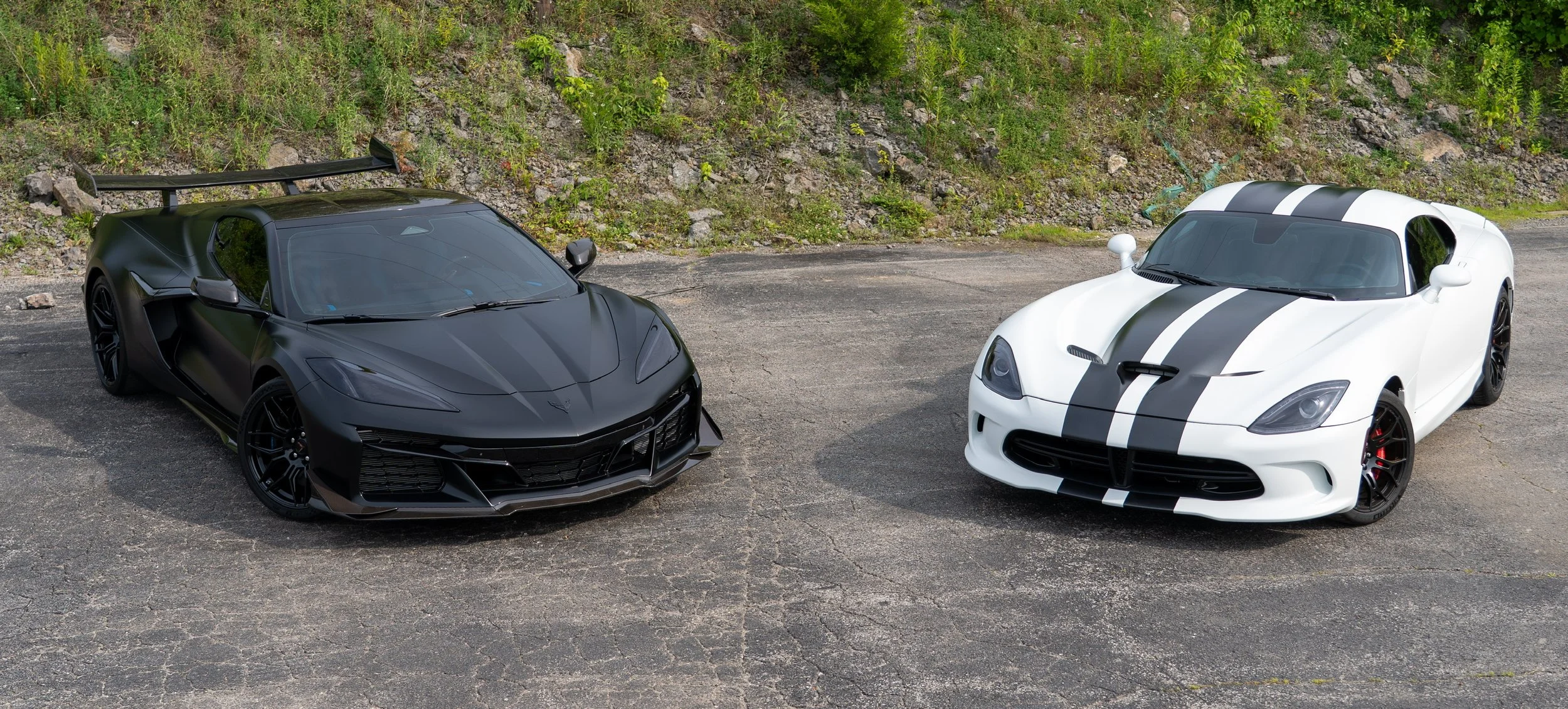 A black sports car with a sleek design and aerodynamic features, and a white sports car with black racing stripes and a bold appearance, are parked side by side on an asphalt surface near a grassy and rocky hillside.