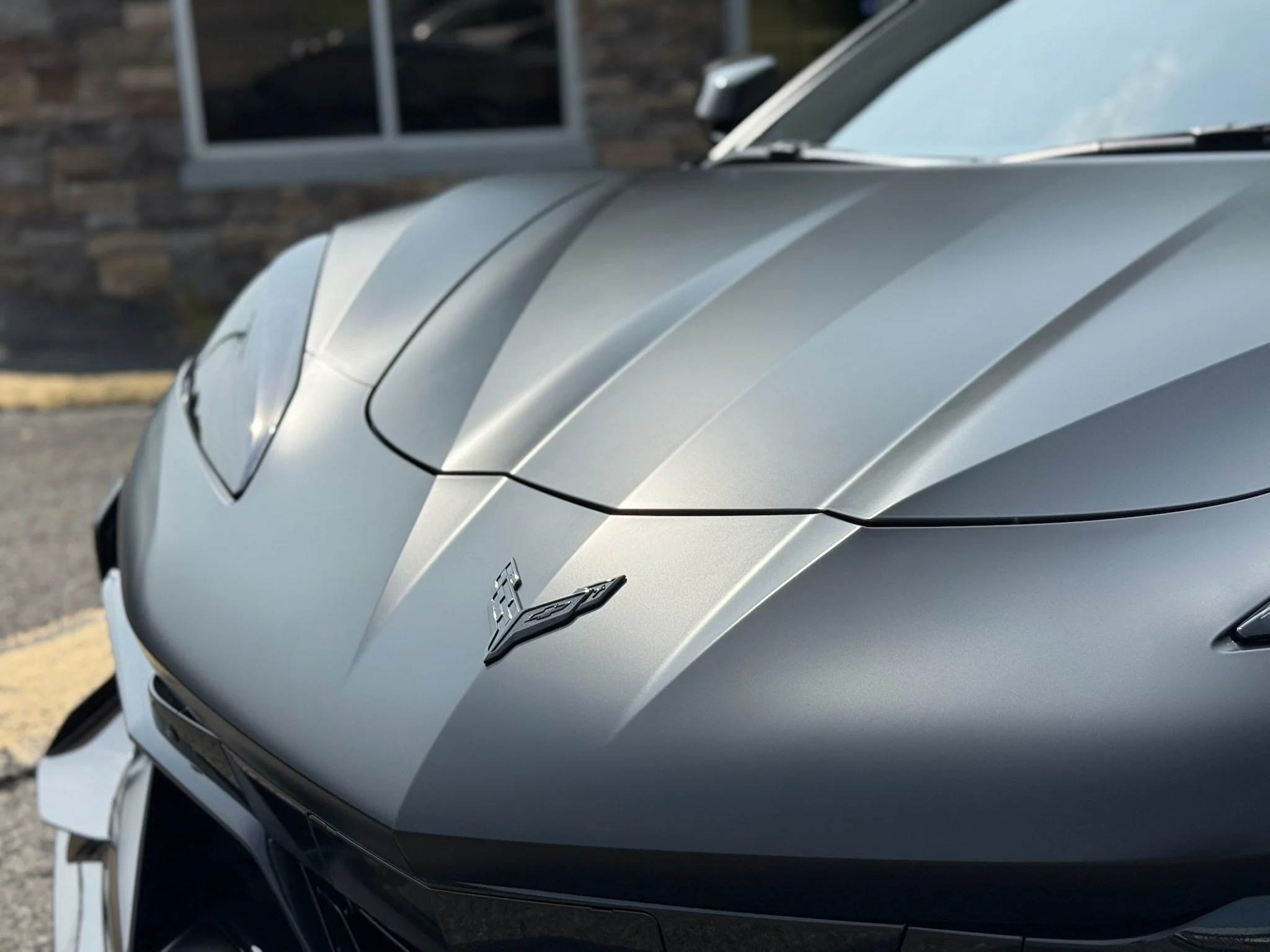 Close-up of the front hood of a silver Chevrolet Corvette sports car, parked outdoors near a building with a window.