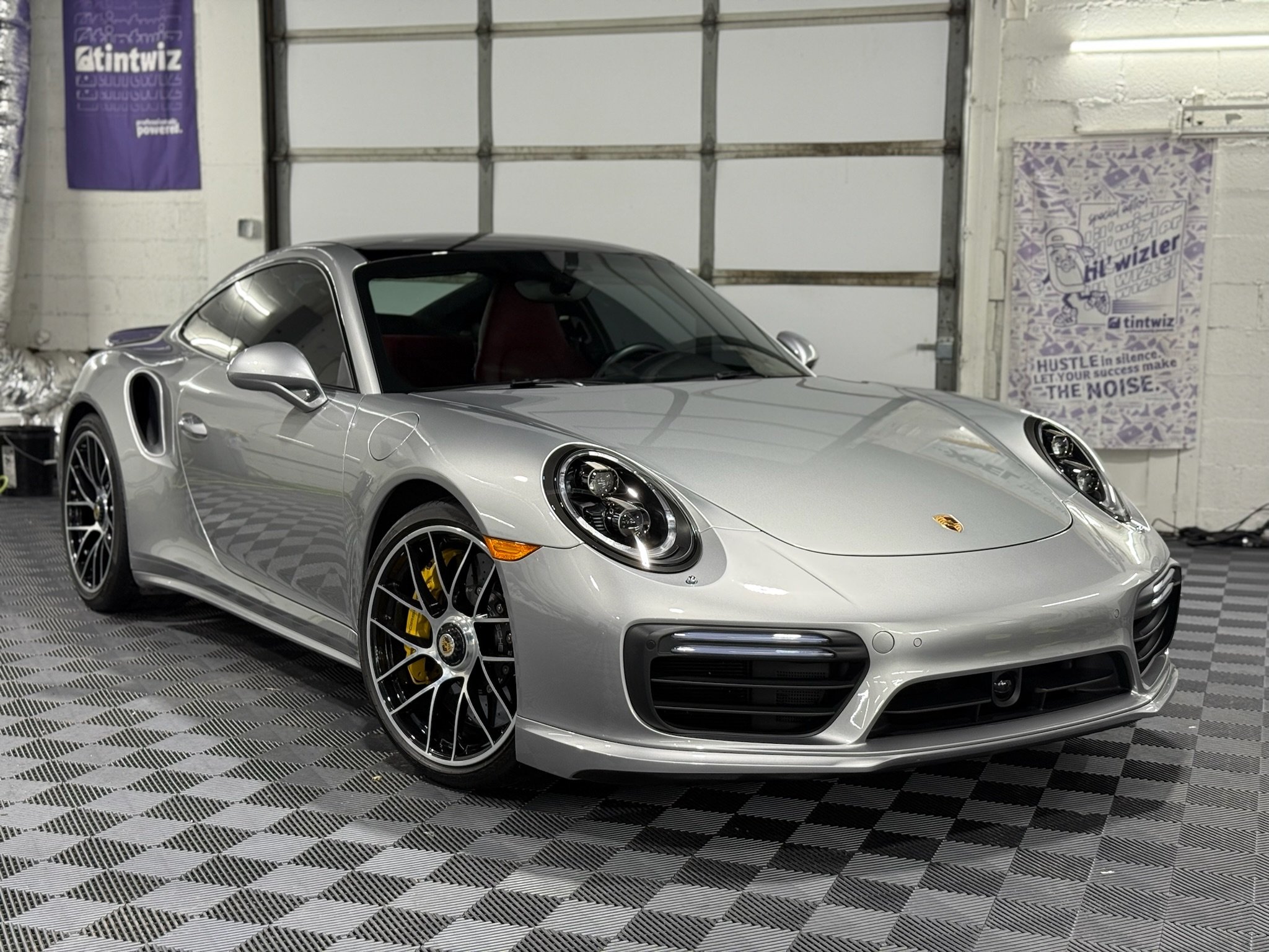 A silver Porsche 911 sports car parked indoors on a checkered floor, with a garage door in the background and purple promotional banners on the wall.