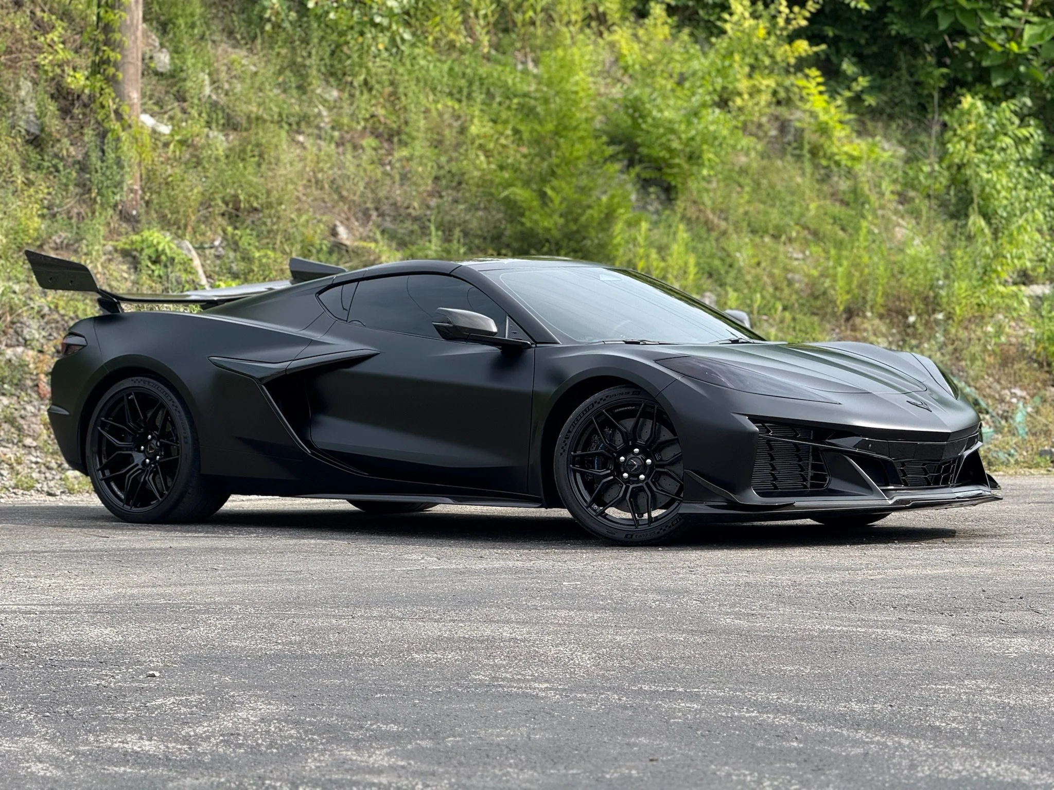 A black sports car with a sleek design, large rear spoiler, and black wheels parked on a paved surface with green foliage in the background.