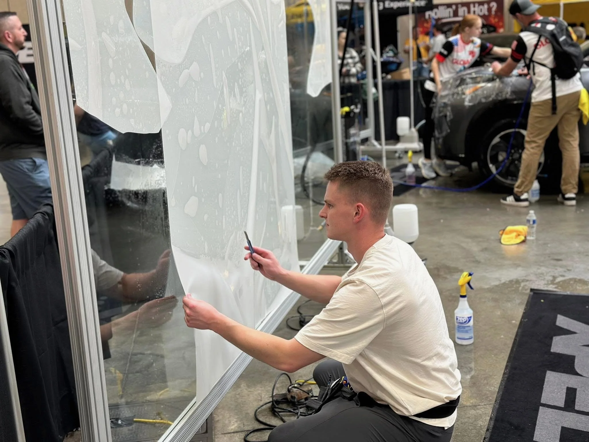 A man is cleaning a glass partition at a car detailing event. In the background, two people are working on a car, and there are cleaning supplies and water bottles on the ground.