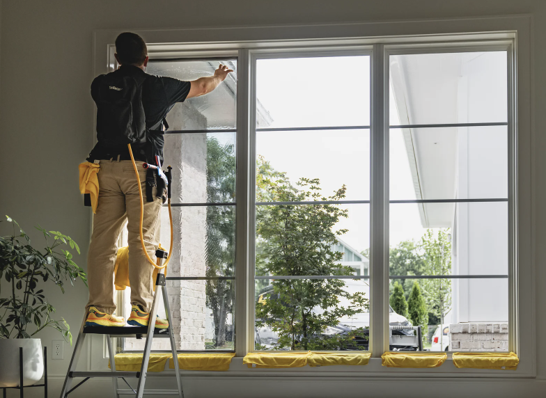 A worker cleaning a large window from the inside of a house, standing on a ladder with cleaning tools and yellow cloths placed on the window sill.