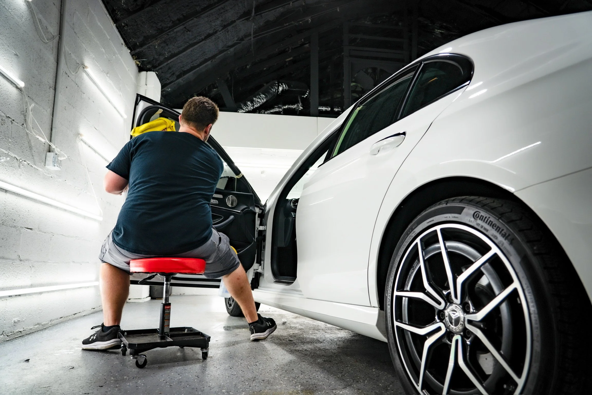 A man repairing a white car inside a garage. The man is seated on a red stool, working on the interior of the vehicle with the passenger door open. The garage has a concrete floor and white brick walls with fluorescent lighting.