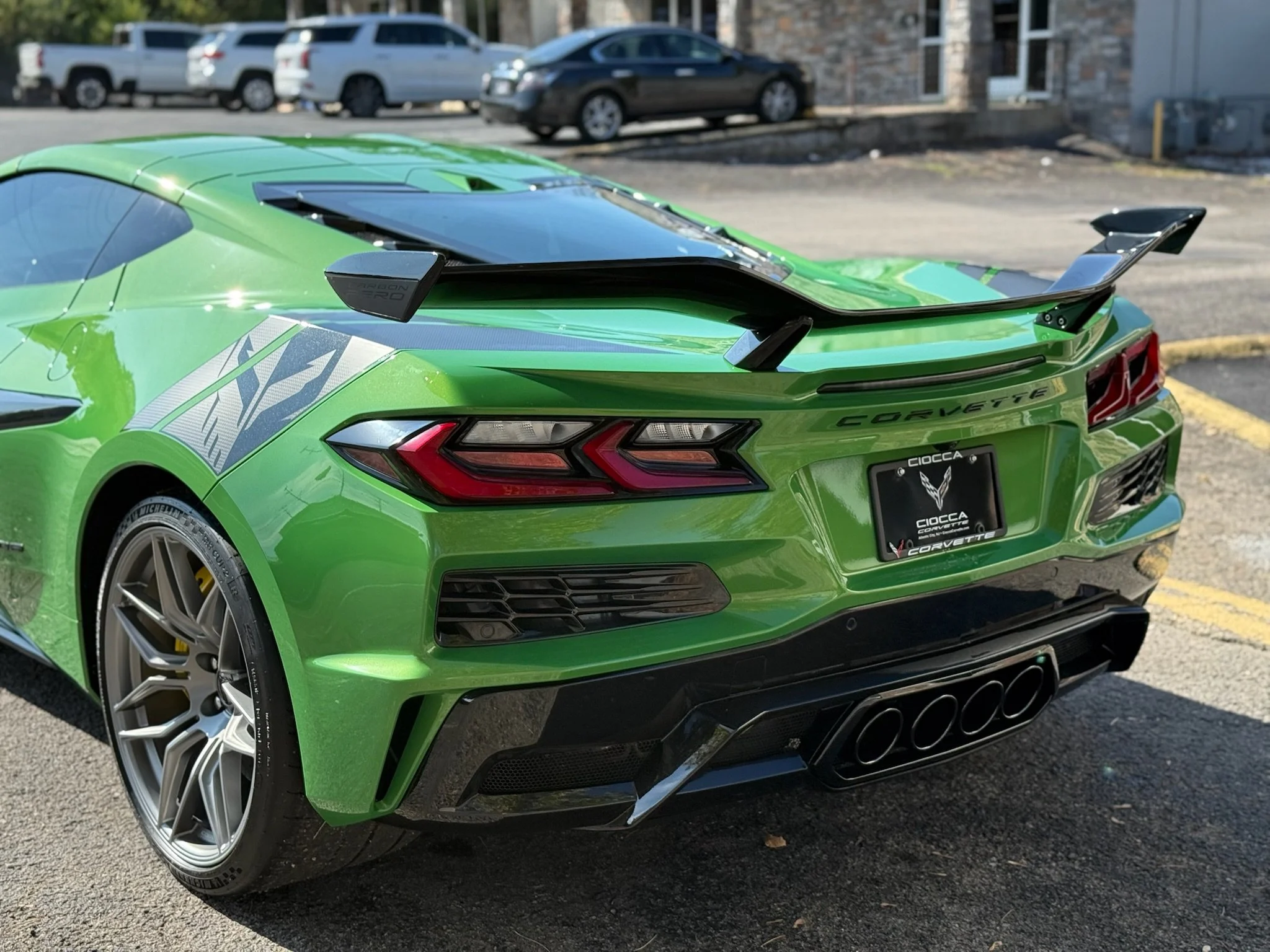 A bright green Chevrolet Corvette parked in a parking lot, showing its rear end with a large spoiler, quad exhaust pipes, and distinctive taillights.