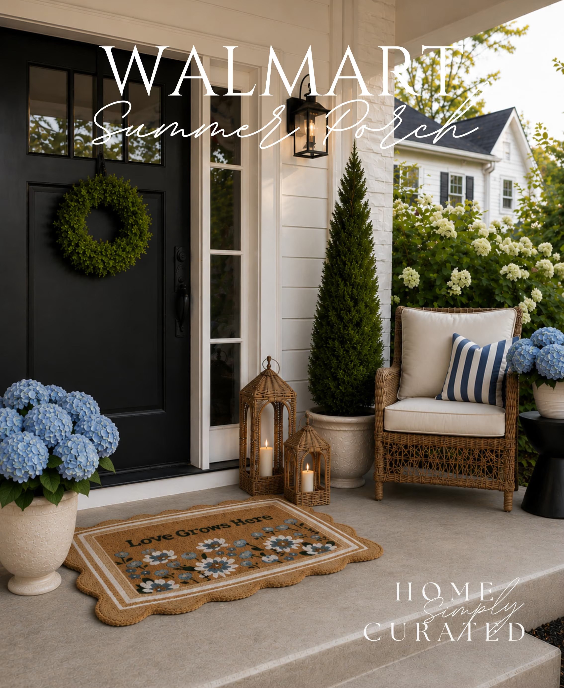 Front porch area decorated with a black door with a wreath, potted blue hydrangeas, a beige doormat with flower patterns and the words 'Love Grows Here', wicker lanterns with candles, a white armchair with striped pillow, a potted evergreen, and white flowering bushes in the background.