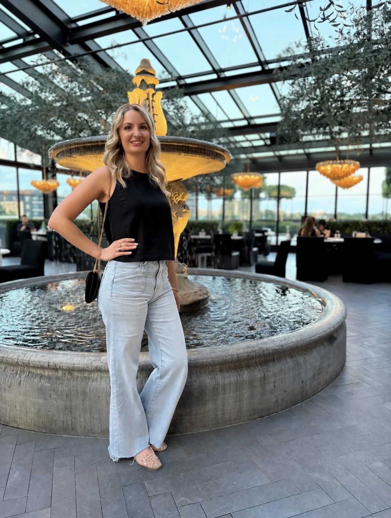 A woman with blonde hair wearing a black sleeveless top and light jeans, standing in front of a decorative fountain inside a glass-enclosed restaurant or cafe with chandeliers hanging from the ceiling.