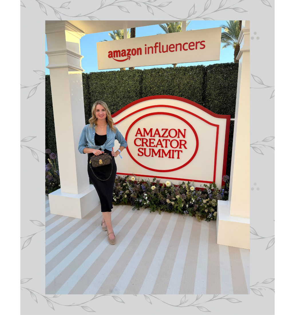 A woman standing in front of a sign for the Amazon Creator Summit at an outdoor event, with a hedge and palm trees in the background.