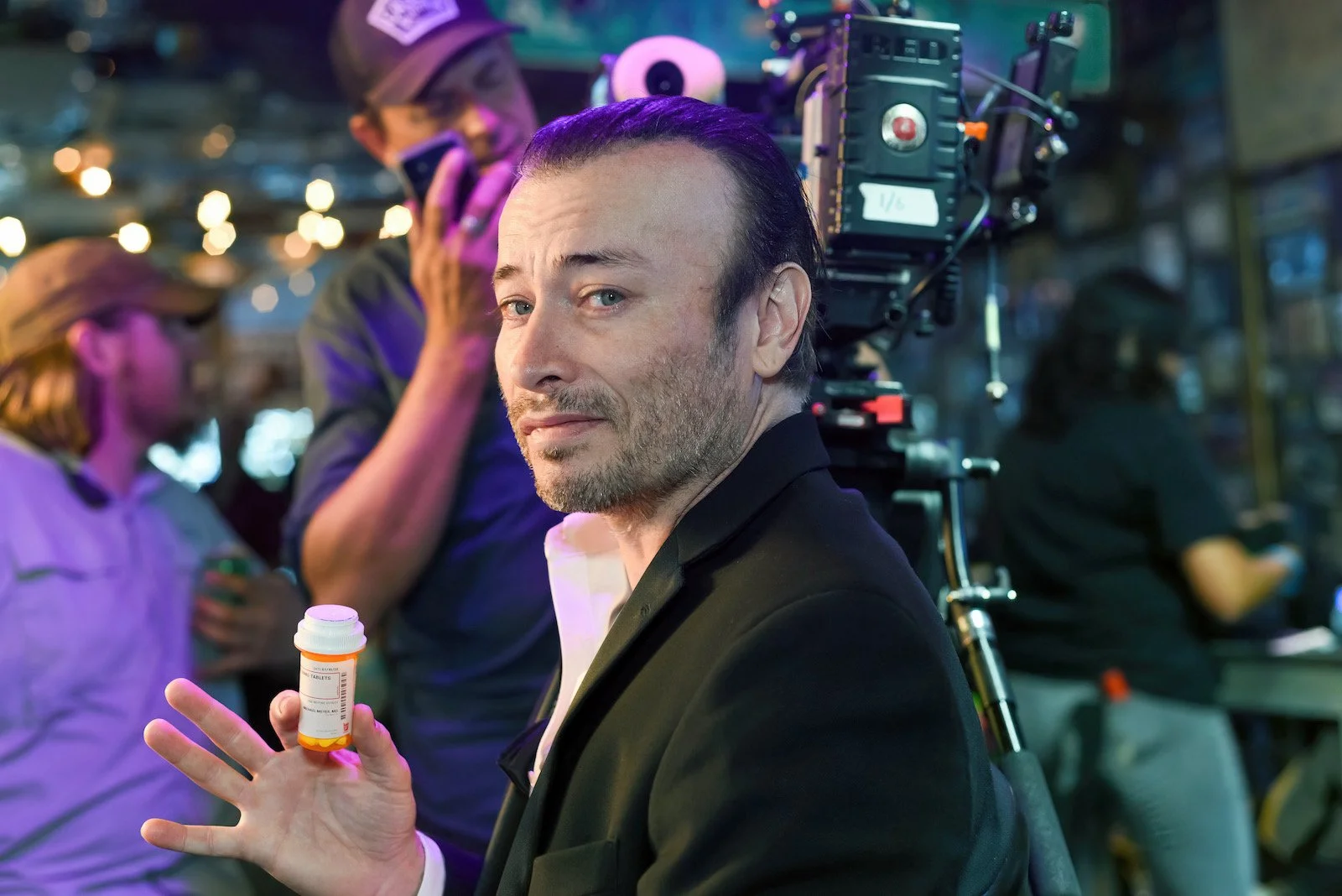 A man with dark hair and a beard, wearing a black blazer, holds a pill bottle in his left hand. Behind him, there are people and camera equipment, suggesting a film or photo shoot setting.