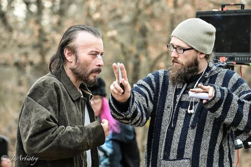 Two men on a film set, one with a beard and glasses, the other with long hair and a beard, engaged in a conversation or disagreement.