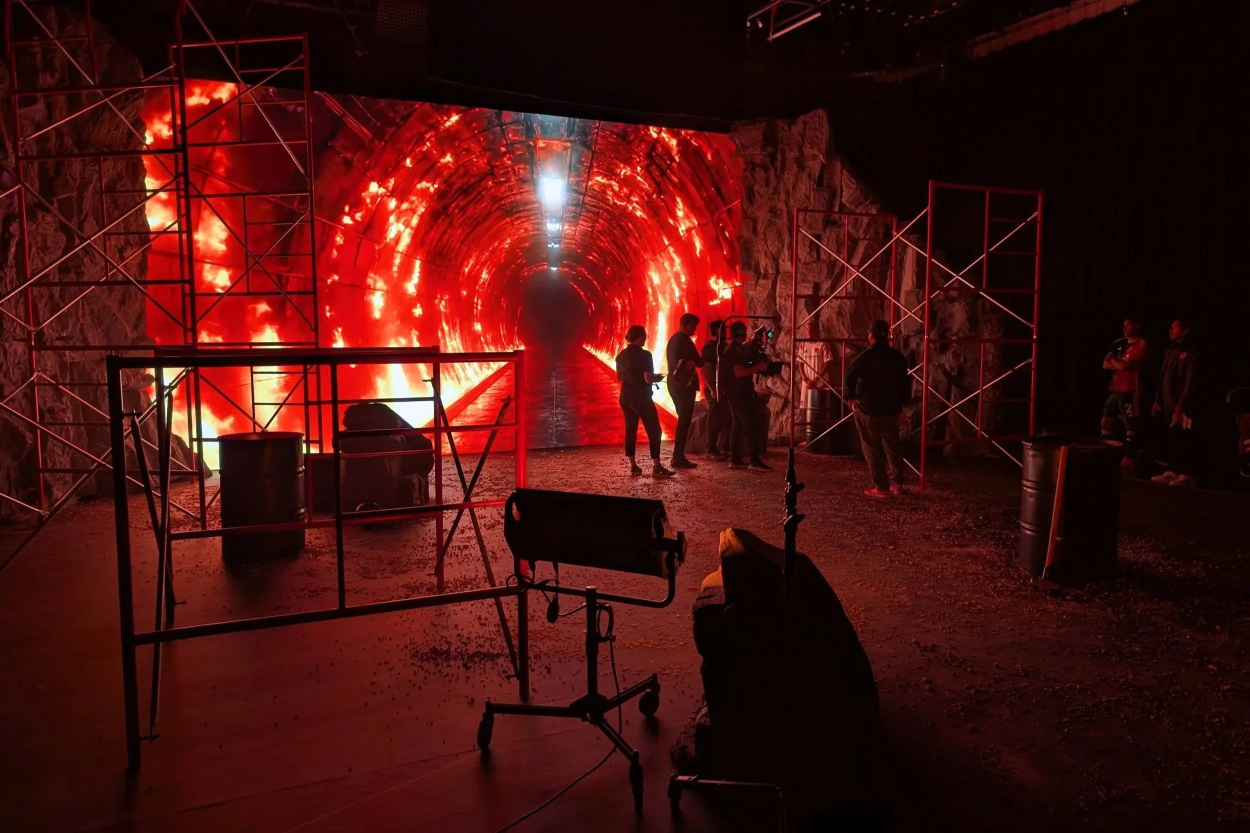 A group of people standing in front of a bright red and orange tunnel with fire effects on a large digital screen, surrounded by scaffolding and equipment, in a dark indoor setting.