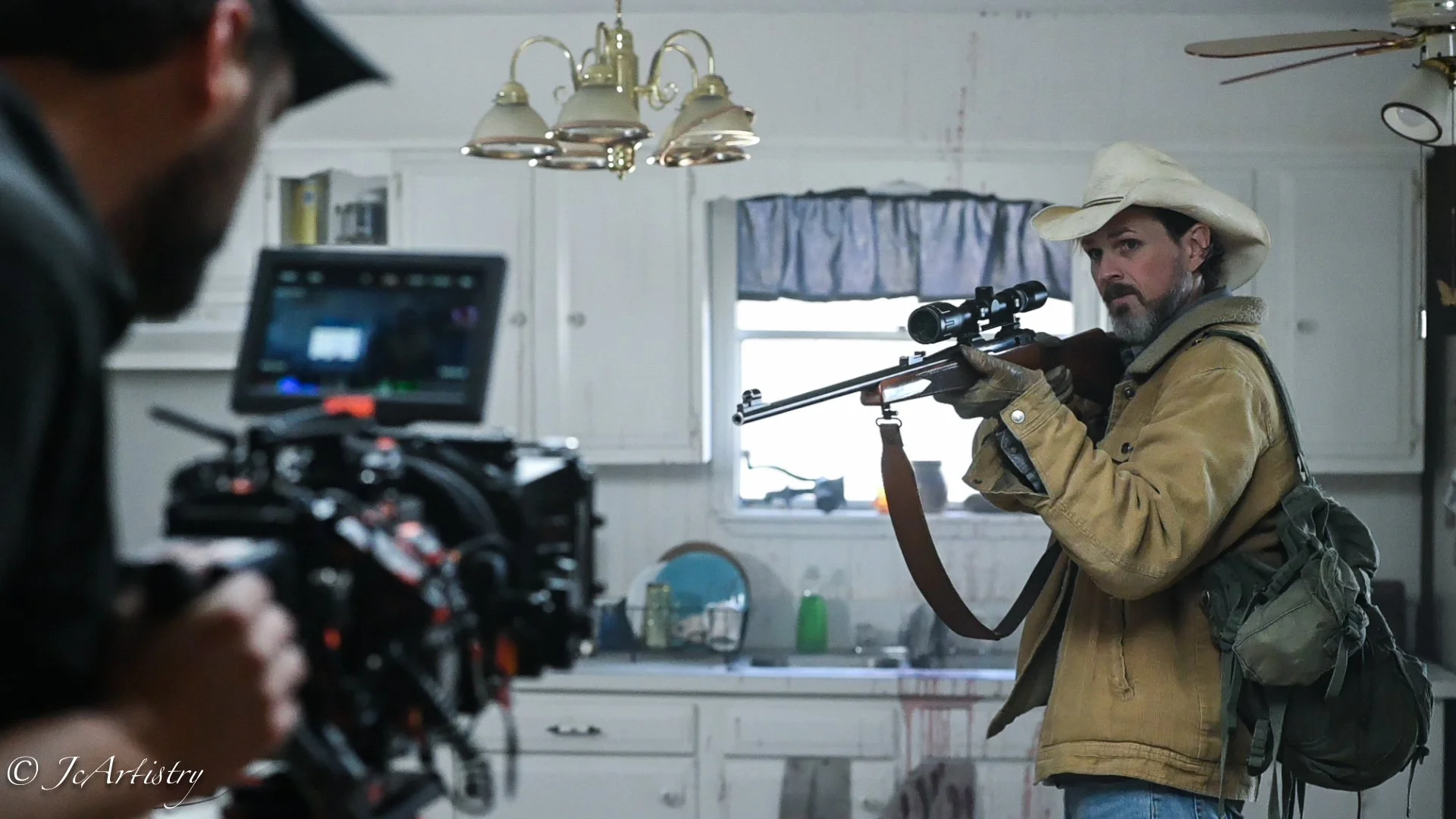 Man dressed as a cowboy aiming a rifle indoors, with a camera operator filming him. The room has white cabinets, a window with a blue curtain, and hanging lights.