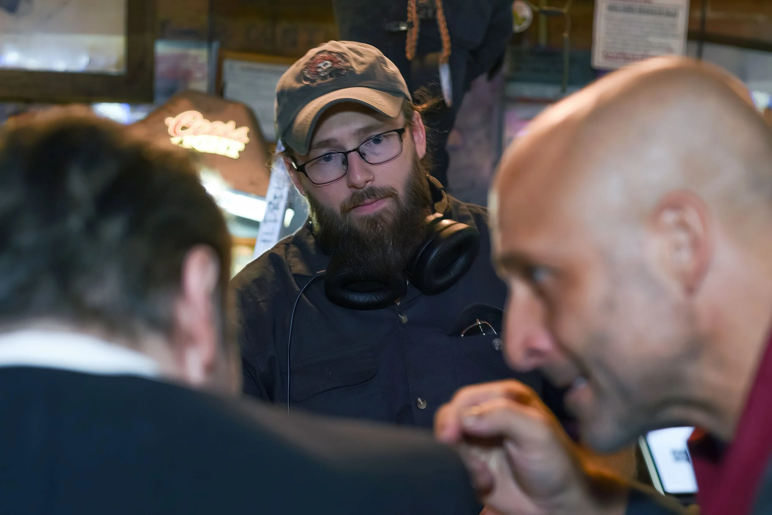 A man with glasses and a beard, wearing a cap and headphones around his neck, is looking at something with other people around him in a crowded indoor setting.
