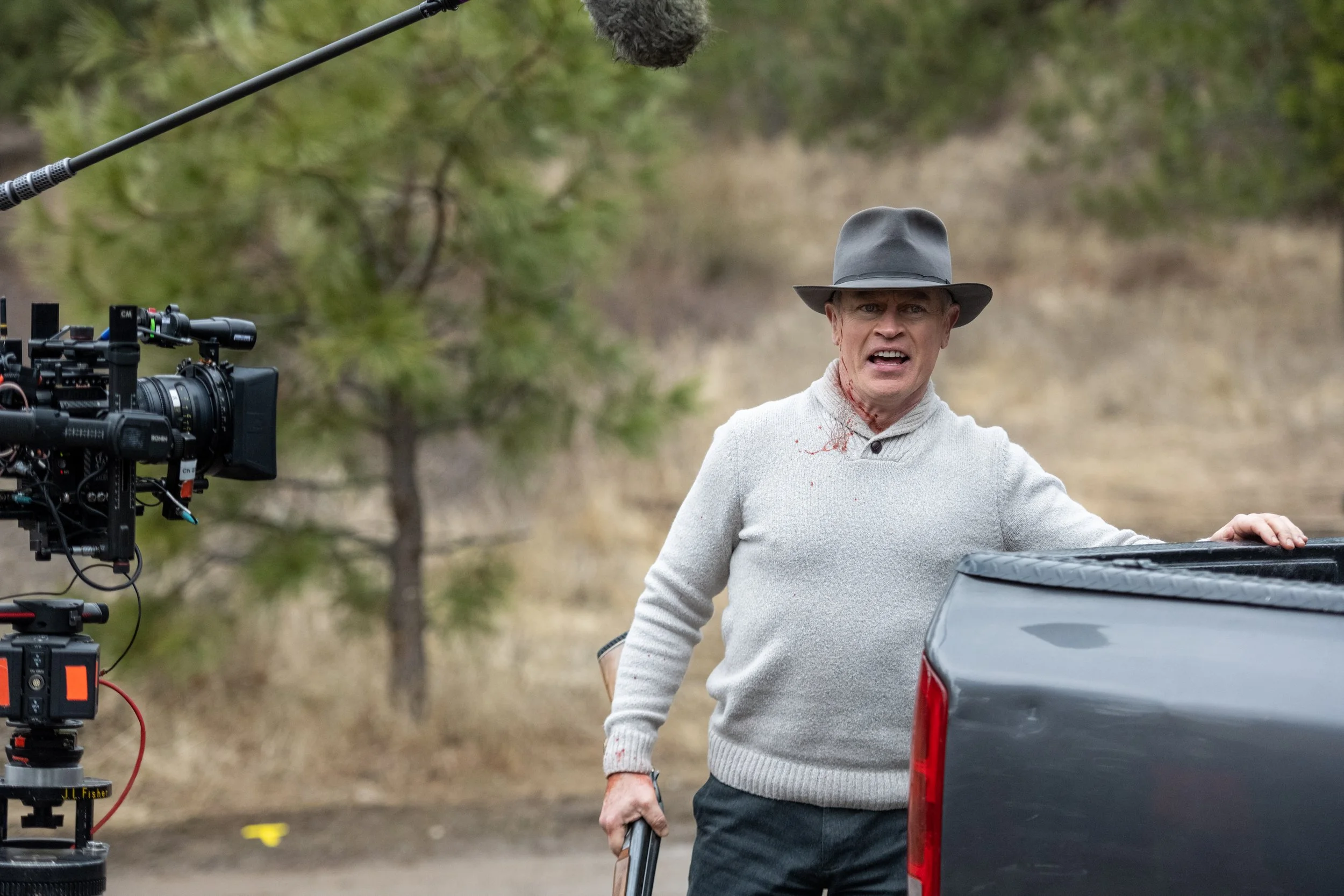 A man in a gray hat and sweater with blood on his neck stands outdoors next to a car, with a film camera and boom microphone filming him.