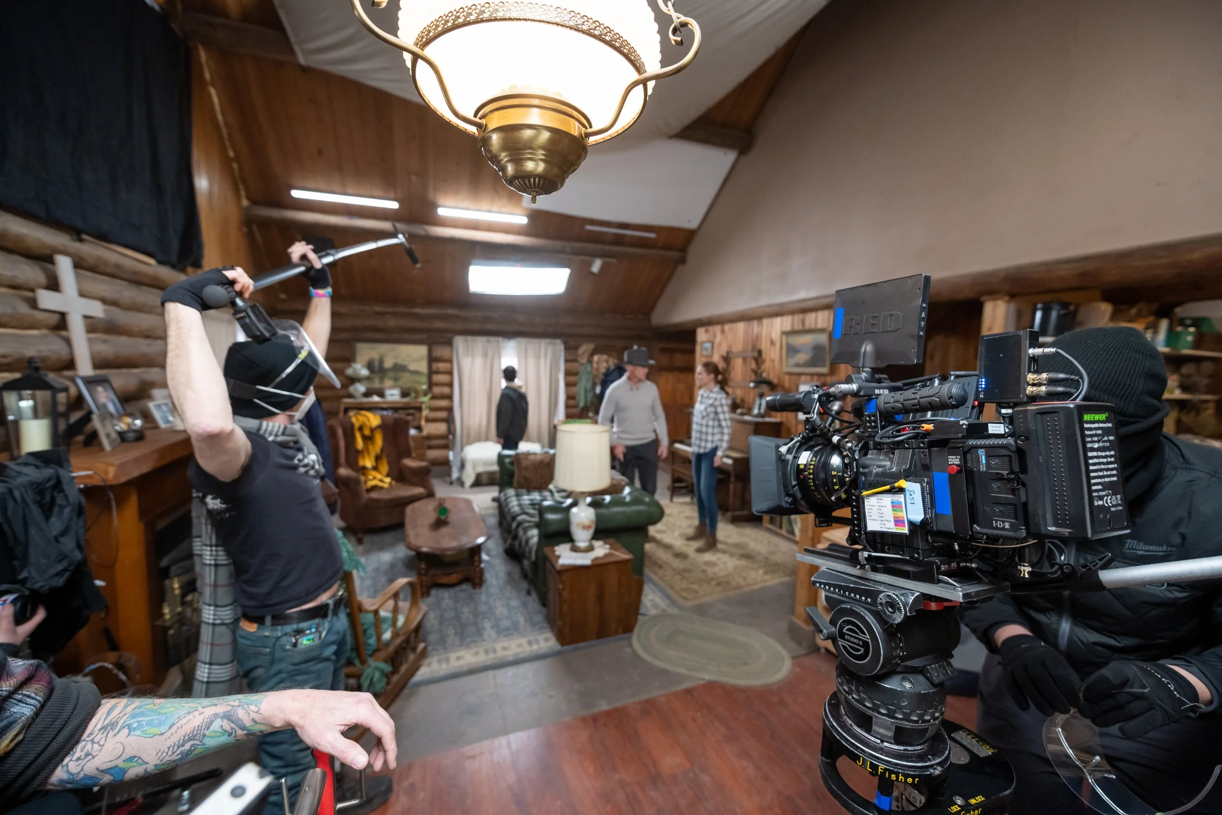 Film crew inside a log cabin with wooden walls and furniture, filming a scene with people in casual and vintage clothing, a woman in a hat, and a man in a gray sweater. Professional camera equipment and crew members are visible.