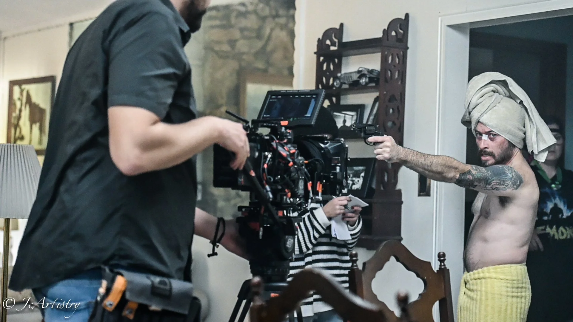 A shirtless man with a towel wrapped around his head and yellow shorts is pointing a gun at a camera on a film set. A crew member is operating the camera, and a person in a striped shirt is holding a clapboard behind them.