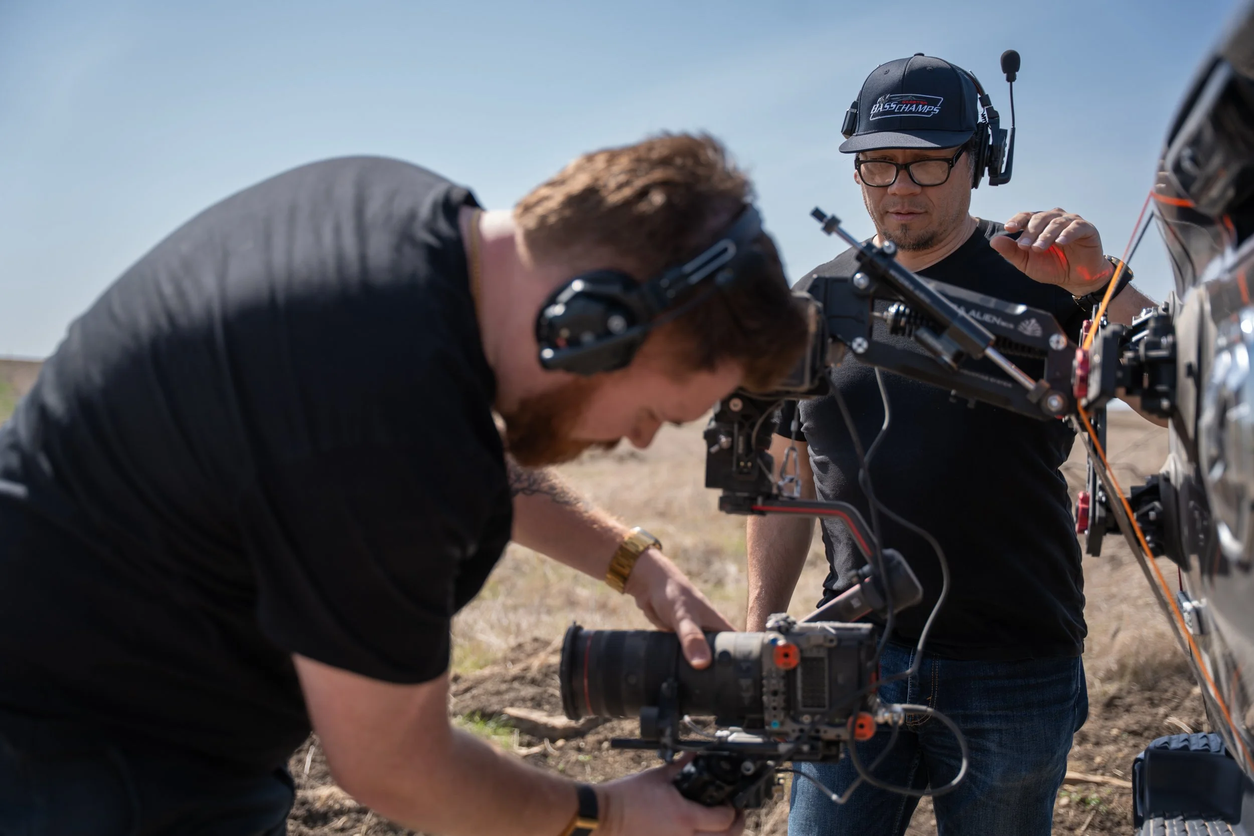 Three men are working outdoors on a drone or helicopter, with one man using a power drill, another adjusting equipment, and the third observing, all wearing casual clothes and headsets.