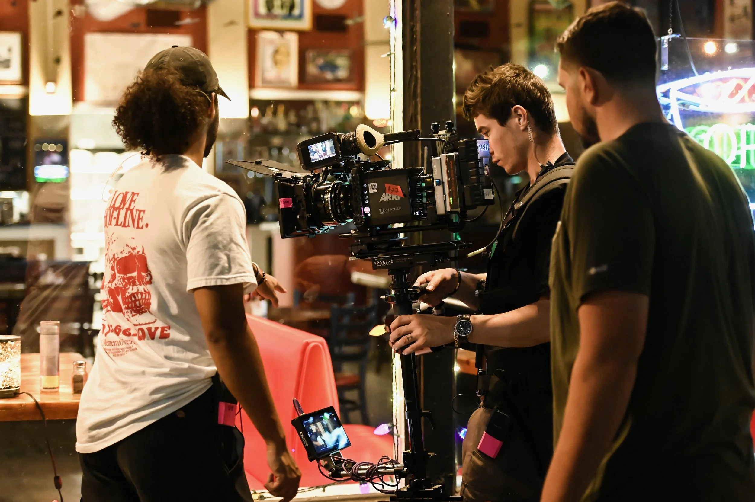 A film crew setting up filming equipment inside a restaurant or bar with neon signs and artwork on the walls. The crew members are gathered around a camera mounted on a tripod, and one person is wearing a white T-shirt with red text.
