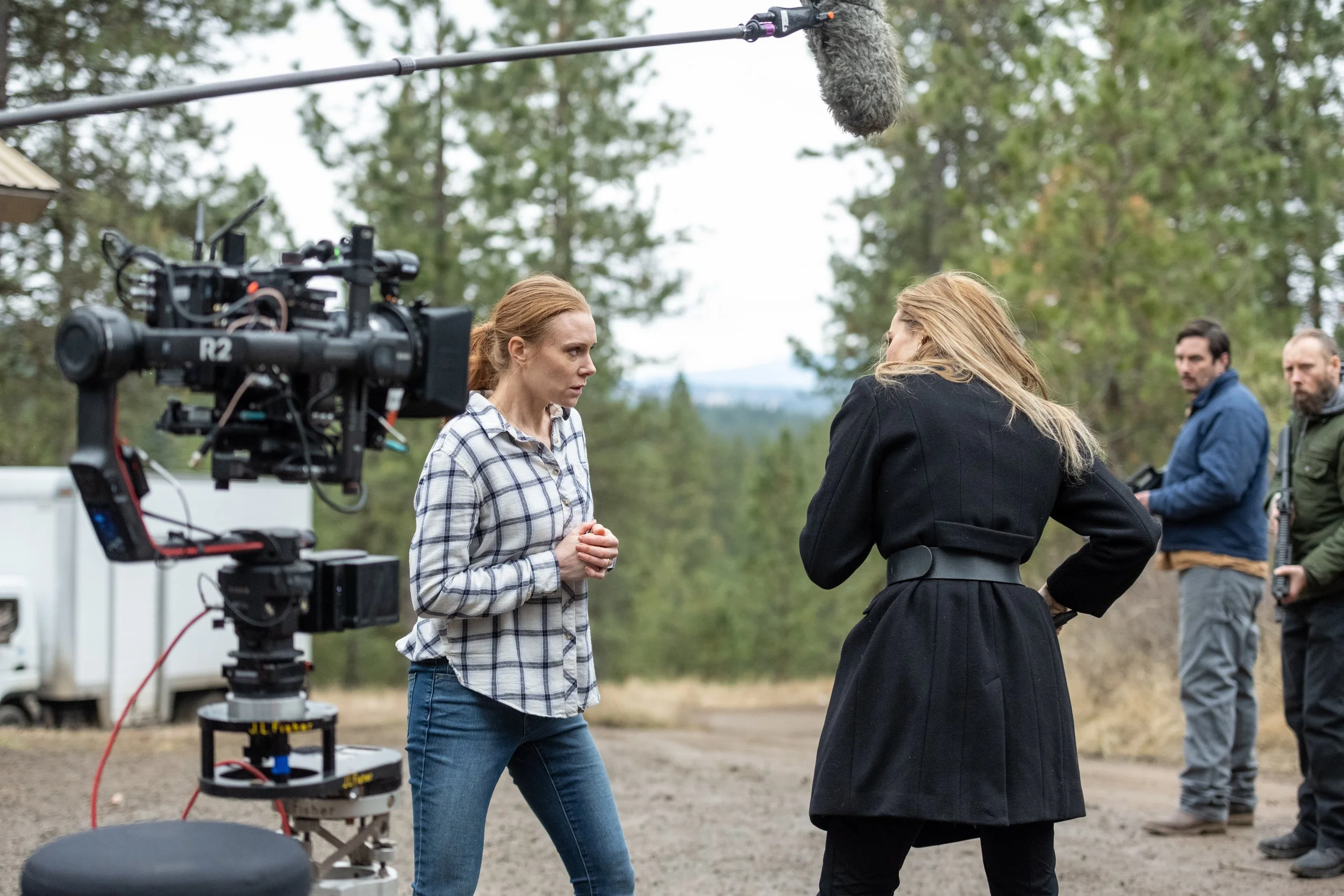 Two women are engaged in a conversation outdoors on a film set, with a film camera and a microphone overhead. In the background, two men are standing and observing.