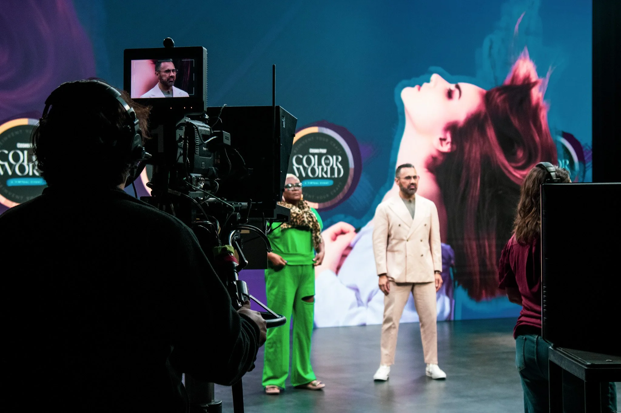 Behind the scenes of the CosmoProf Color the World event showing the hosts on stage, crew members, and a camera operator filming the event, with a large screen displaying a model's image in the background.