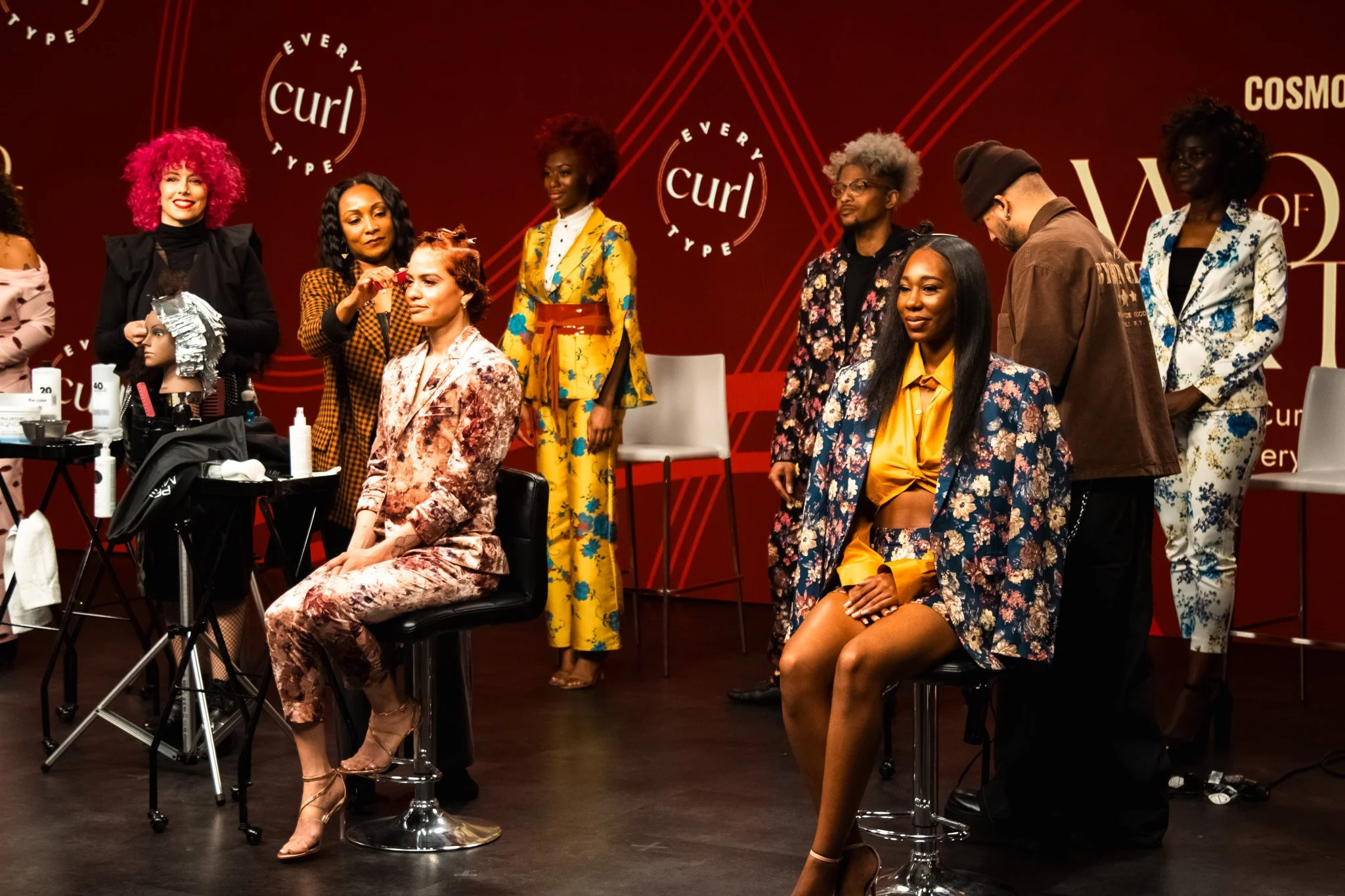 Model sitting in front of a hairstylist with other models standing behind in a salon setting with a red wall displaying the 'Every Curl' logo.